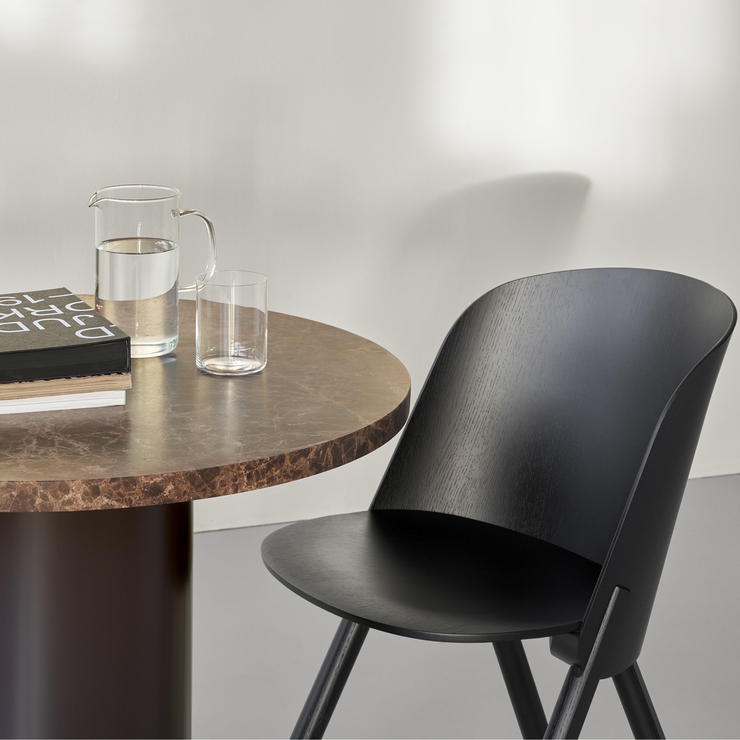 A sleek black chair sits next to the E15 CT09 ENOKI BISTRO table with a rich marble top. Atop the table are a glass pitcher, a glass of water, and a closed black book with white text, against a light-colored wall backdrop.