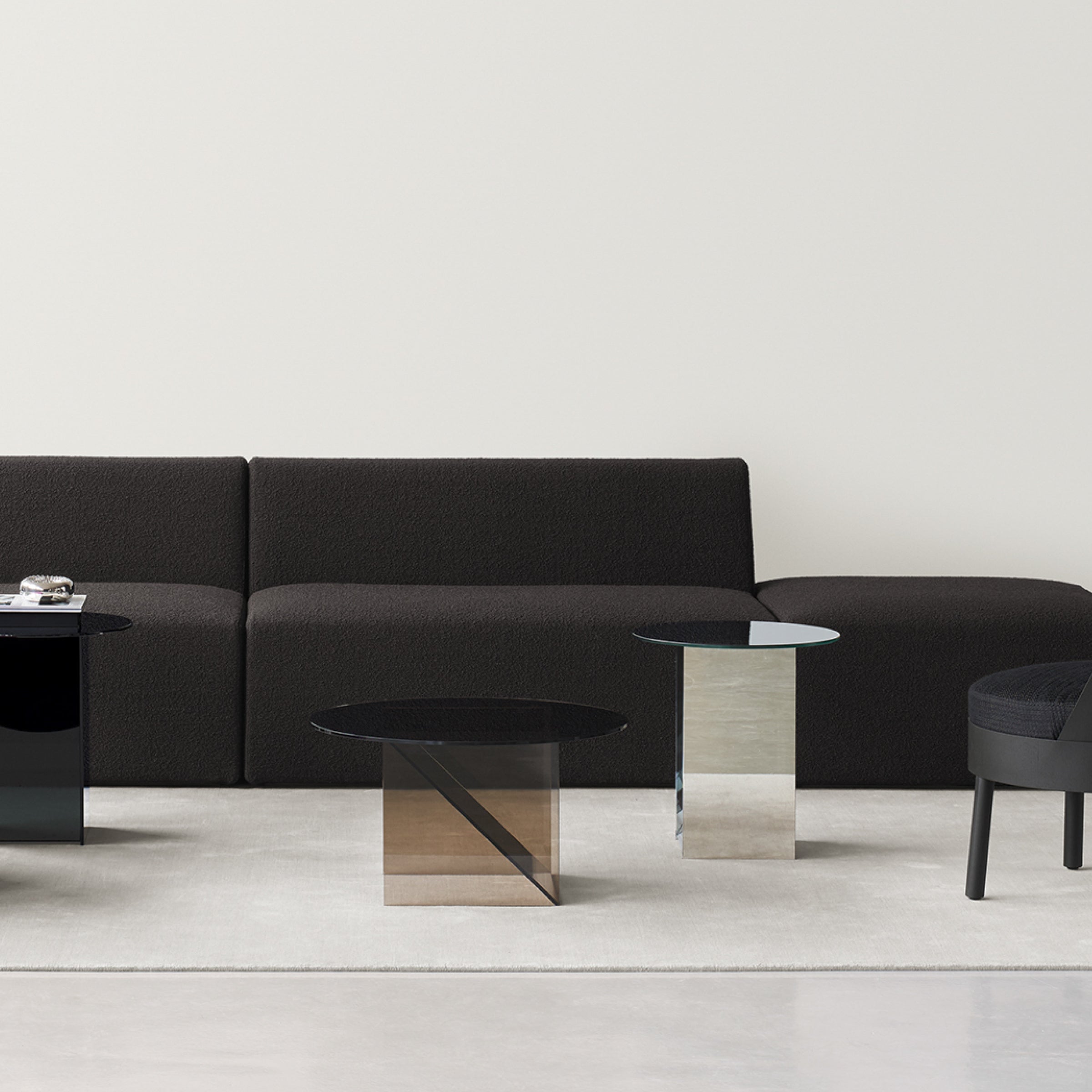 A minimalist living room showcases a black sofa and three E15 CT11 Kaisa side tables that combine sculptural elegance with practicality. A dark upholstered chair to the right enhances the neutral palette of sleek, contemporary furniture arranged on a light rug.