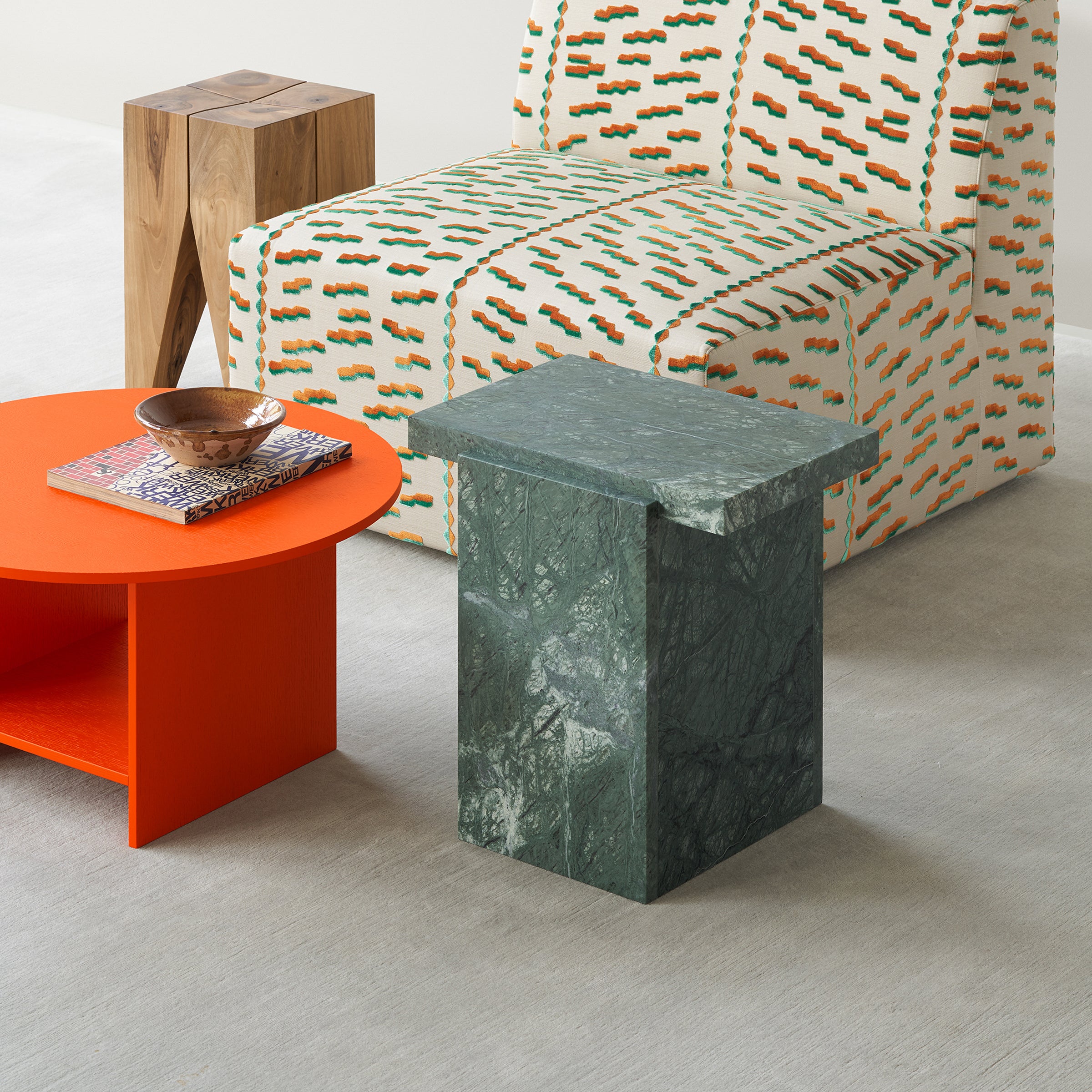 A modern living room showcasing the E15 CT12 Heiji, a geometric green marble side table, and a bright orange round coffee table adorned with books and a wooden bowl. Complemented by a patterned armchair in green and orange, lacquered plywood accents subtly reflect Japanese design principles on a light background.