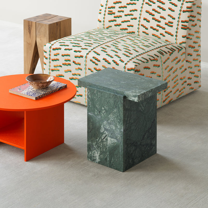 A modern living room showcasing the E15 CT12 Heiji, a geometric green marble side table, and a bright orange round coffee table adorned with books and a wooden bowl. Complemented by a patterned armchair in green and orange, lacquered plywood accents subtly reflect Japanese design principles on a light background.