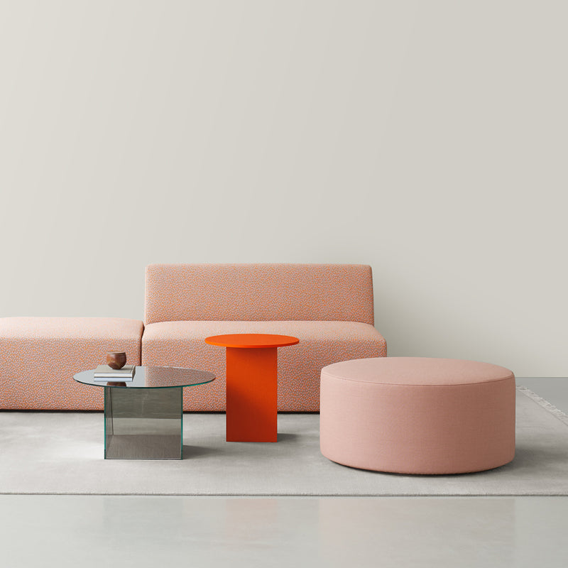 A modern living room features a minimalist orange-patterned sofa, small round pink ottoman, and a round glass coffee table with a square base. These pieces pair seamlessly with the E15 CT12 Heiji side table by Philipp Mainzer, all set against a light-colored wall on a soft rug.