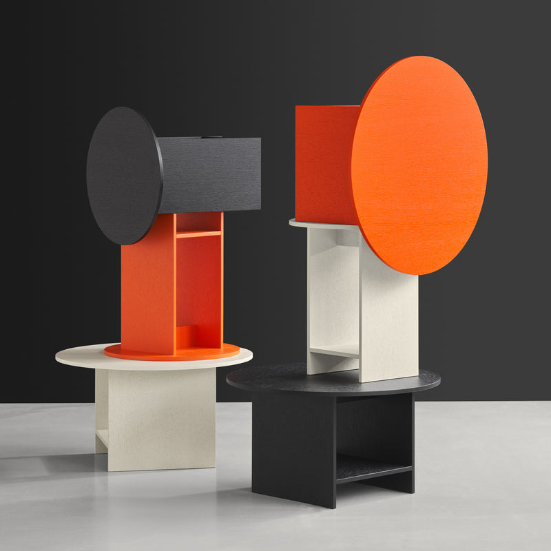 The CT12 Heiji side tables by E15 feature abstract geometric shapes: a black circle and red block on the left, an orange circle with a gray base on the right. These lacquered plywood pieces capture minimalist Japanese design set against a dark background on a light floor.