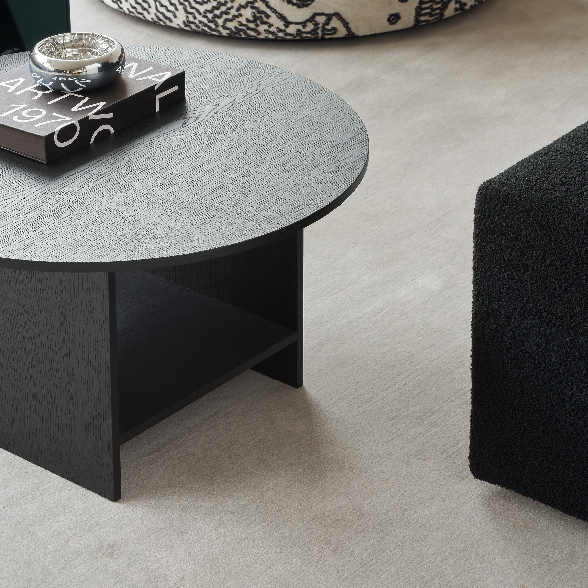 A round, black wooden coffee table with books and a decorative object rests on a beige carpet, accompanied by a textured pouf that adds to the modern minimalist vibe akin to Philipp Mainzers design. This setup provides an alternative to E15s CT12 Heiji side table.