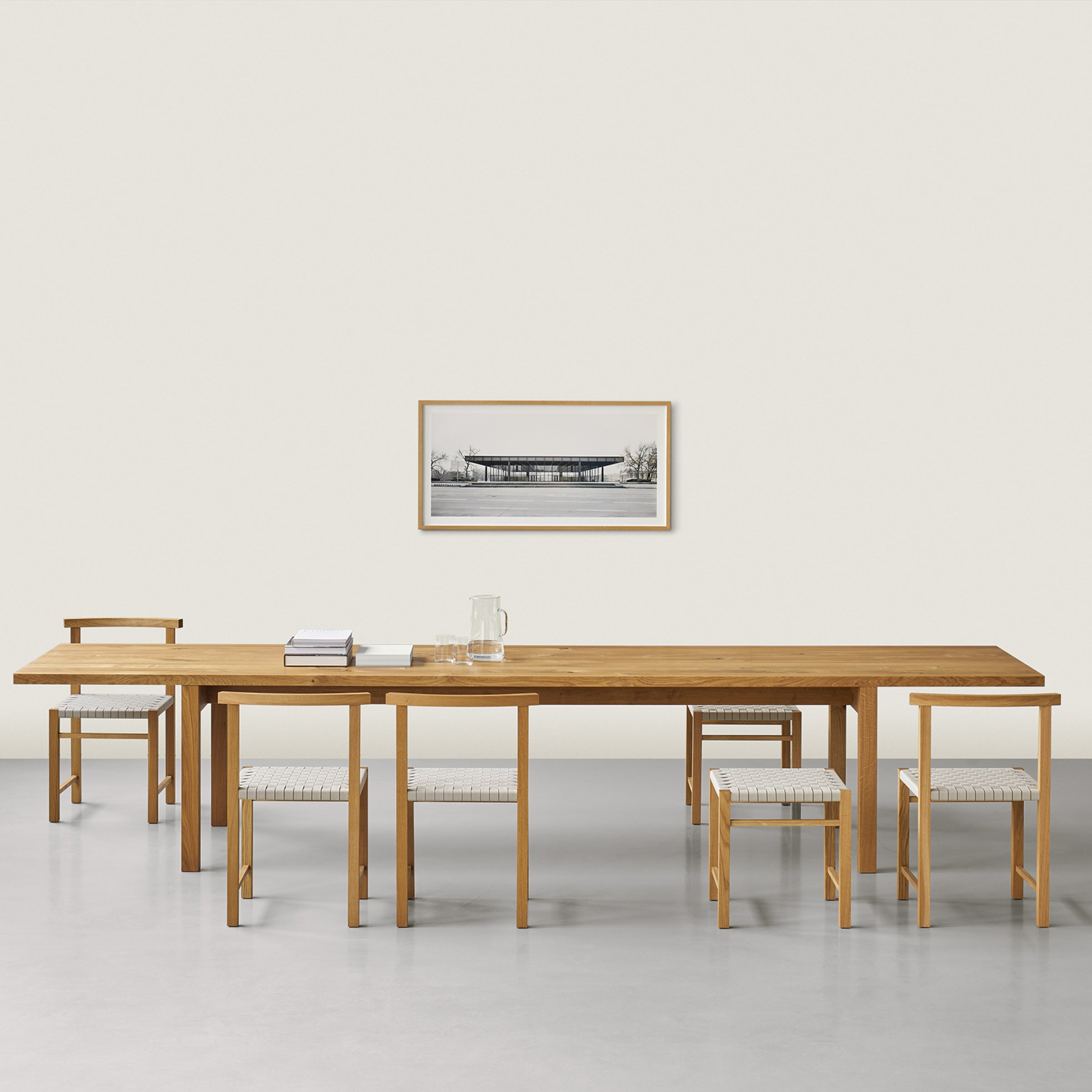 A minimalist dining room showcases E15s DC09 Galerie, featuring a long solid oak table, four chairs, and a bench with light cushions. A black-and-white landscape photo is framed on the light wall above a floor with a light gray hue.