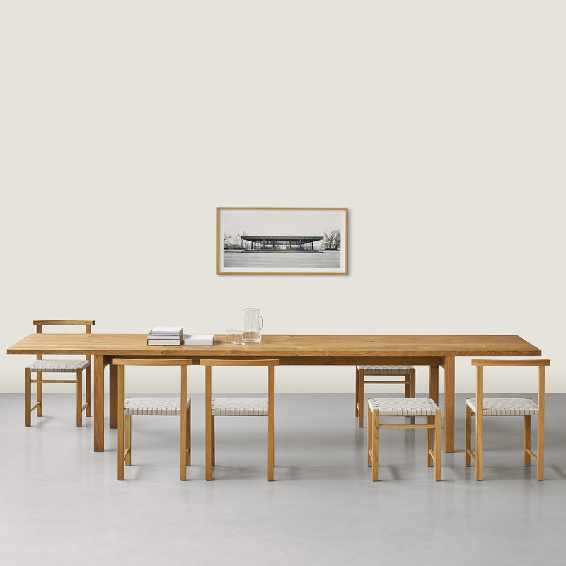 A minimalist dining room showcases E15s DC09 Galerie, featuring a long solid oak table, four chairs, and a bench with light cushions. A black-and-white landscape photo is framed on the light wall above a floor with a light gray hue.