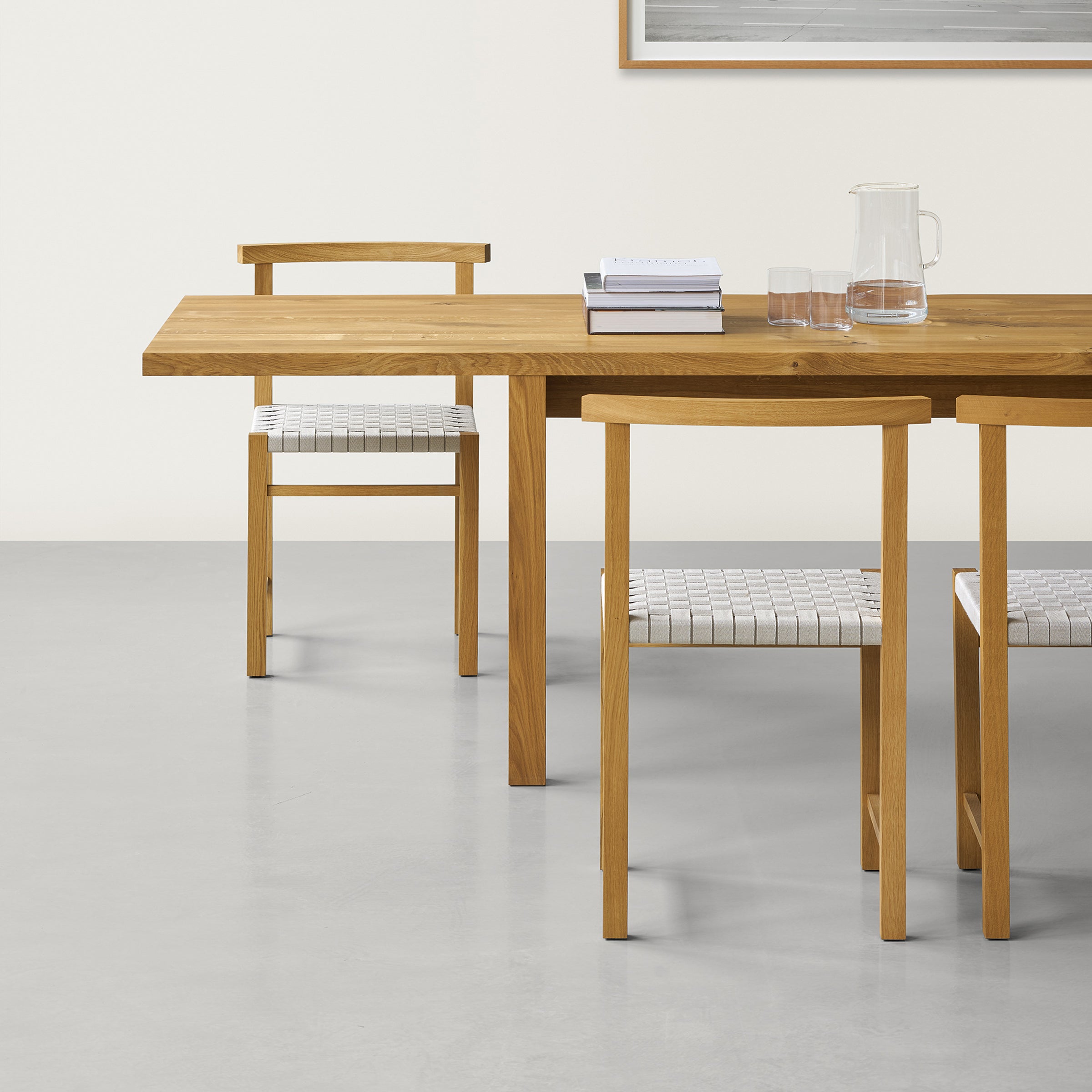 In a minimalist dining room, an E15 solid oak table with two matching chairs is adorned with a stack of books, a glass pitcher, and two glasses. The backdrop features a light-colored wall with framed artwork reminiscent of David Chipperfields DC09 Galerie design.