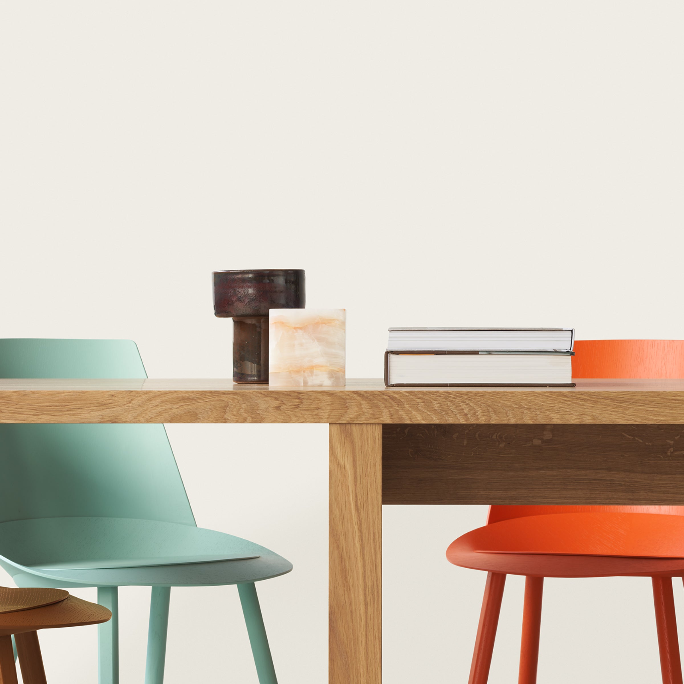 In a minimalist indoor setting, an E15 DC09 Galerie table by David Chipperfield is adorned with books, a stone sculpture, and a cylindrical object. Light blue and orange chairs are tucked under the solid oak table against a plain white wall in the background.