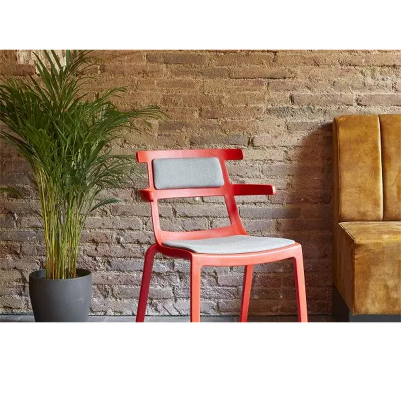 The Resol Tokyo Chair Upholstered, featuring a red frame and light gray cushions designed for UV protection, sits beside a potted plant and brown cushioned bench. The setup is adaptable for interior or exterior use, enhanced by a textured brick wall backdrop.