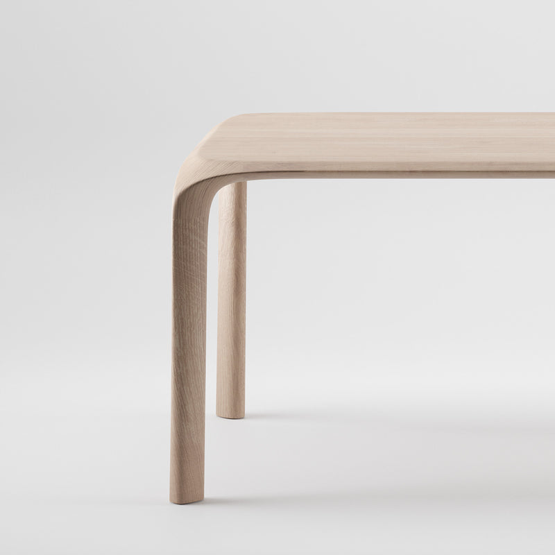 The Artisan Ging Table is a minimalist design crafted from solid wood. Its smooth, curved corners and legs are shown against a plain white background, while the light natural finish enhances its sleek, modern look by highlighting the organic beauty of the wood.