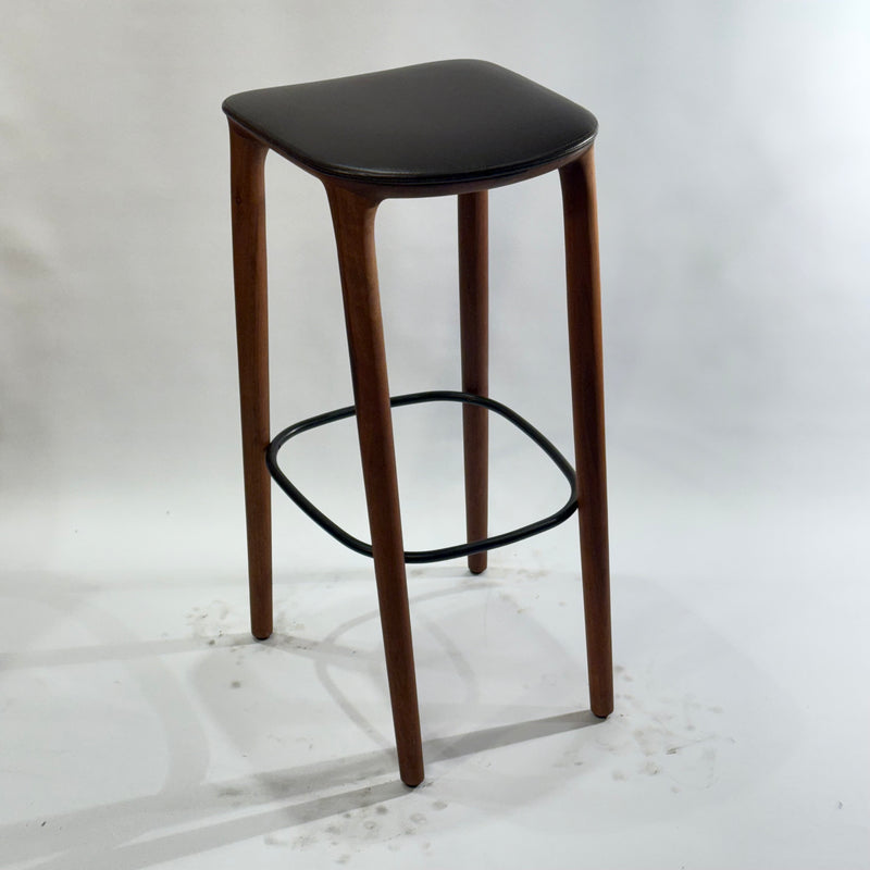 Tall bar stool with dark cushioned seat, slender curved wooden legs, and oval metal footrest on white background