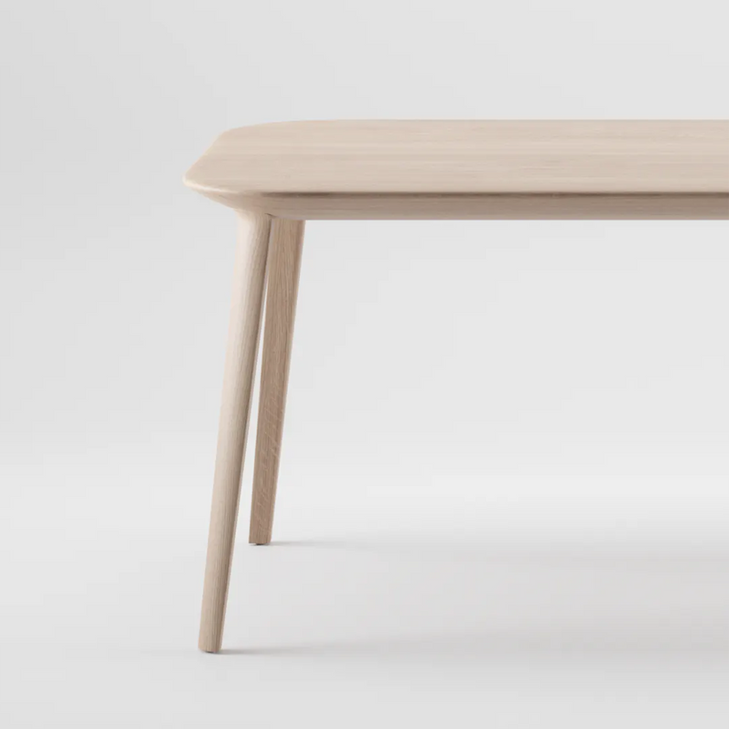 The KALOTA Table by Artisan is a minimalist wooden table with a smooth, light finish and natural oils. It features rounded edges, four angled legs, and its simple yet elegant design is showcased against a plain white background from a corner view.