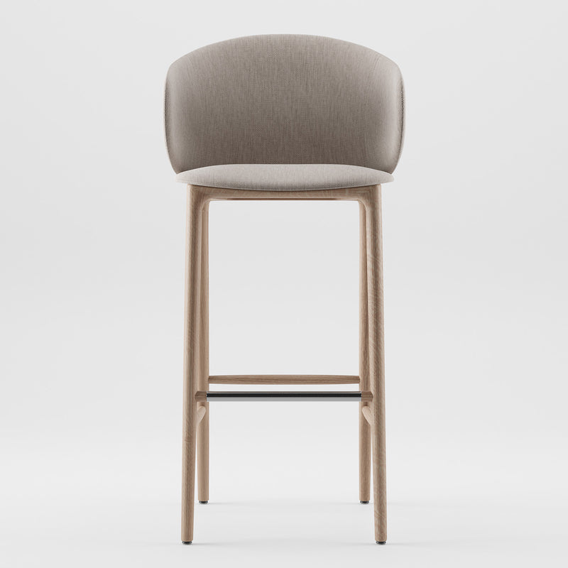 The Artisan Mela Bar Chair is a modern stool with a beige upholstered shell and curved backrest. It showcases mid-century inspiration with light wood legs and a sleek black metal footrest, all against a plain white backdrop.