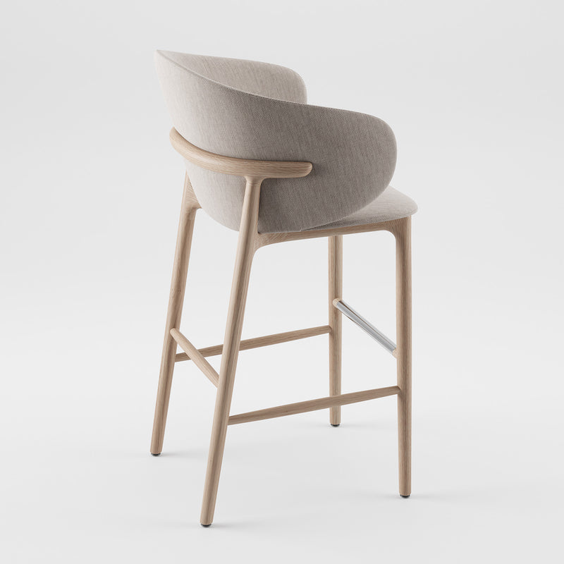 The Artisan Mela Bar Chair offers a mid-century modern vibe with a curved upholstered backrest and seat in light gray fabric, complemented by a natural wood finish and sleek metal footrest, set against a plain white background.