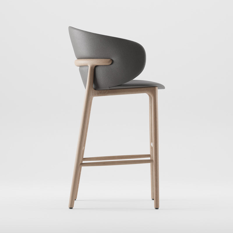Side view of the Mela Bar Chair by Artisan, showcasing a mid-century style with a gray upholstered curved seat and backrest. The chair’s minimalist aesthetics are complemented by subtle curves and clean lines on its light wooden frame.