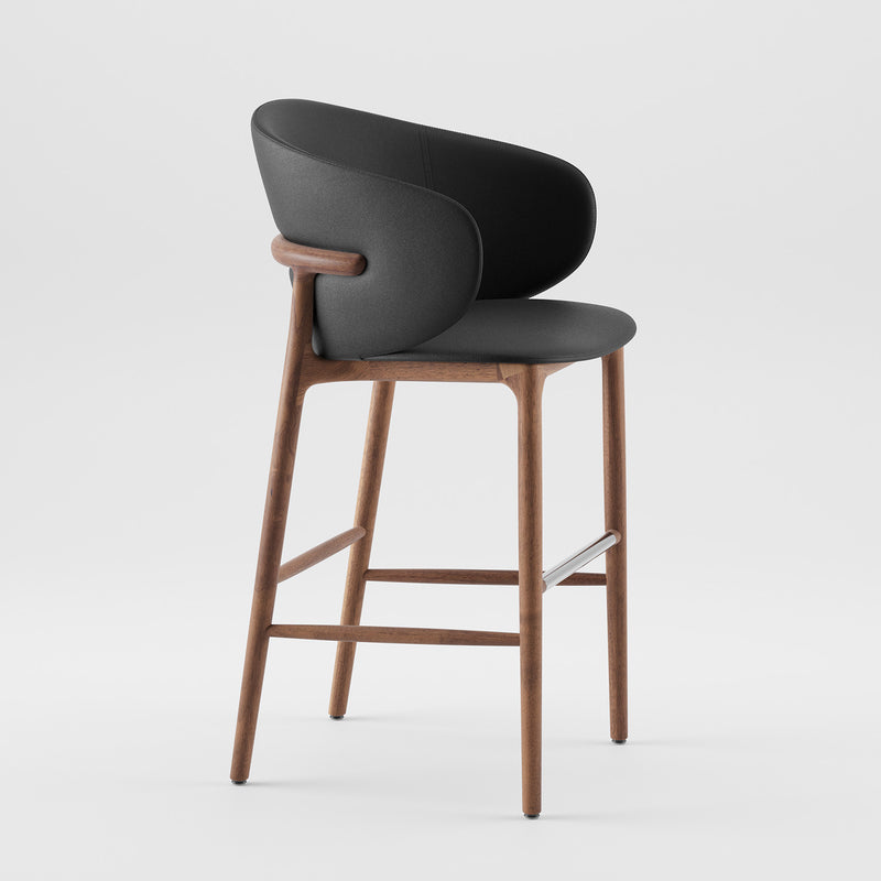 The Artisan Mela Bar Chair combines a sleek wooden frame with a black upholstered backrest. Its mid-century design has clean lines and a metal footrest, standing elegantly on a plain white background.