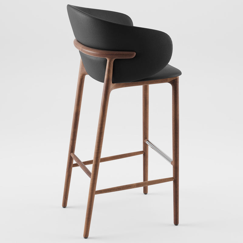 The Artisan Mela Bar Chair, with its sleek black seat and curved wooden backrest, evokes mid-century design. Complemented by wooden legs and a metal footrest, it delivers a modern minimalistic look against a white backdrop.