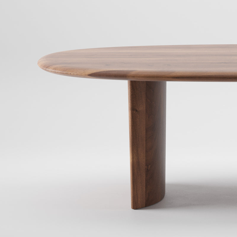 Close-up of the Artisan MONUMENT Table, a rounded wooden table with a solid wood, smooth polished finish enhanced by natural oils. Its visible grain enriches the texture and sustainable minimalist design. A sturdy cylindrical leg stands against a plain white background.