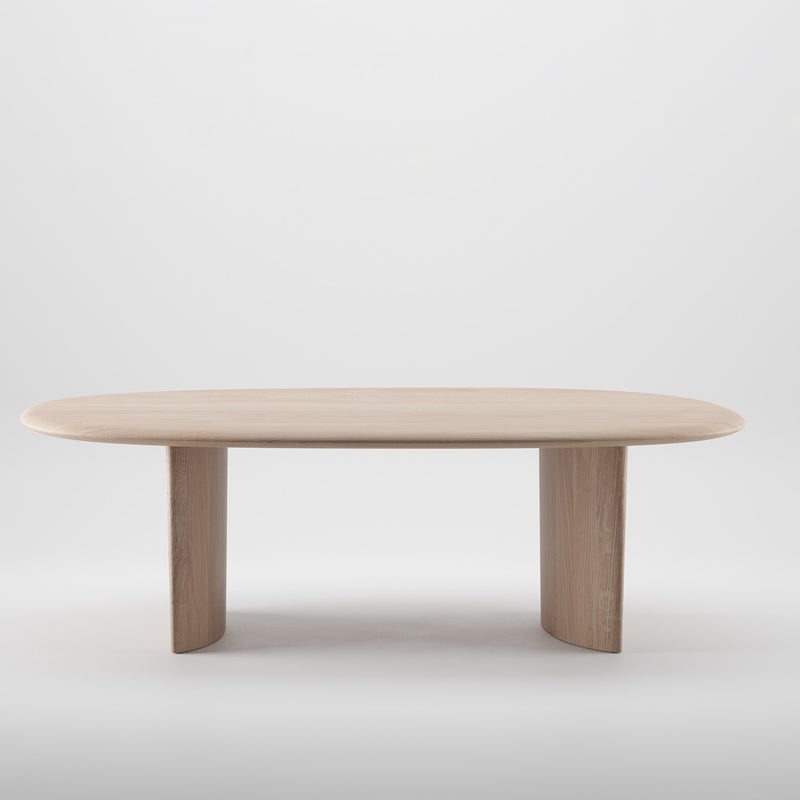 The MONUMENT Table by Artisan is a minimalist, oval-shaped solid wood table with a light finish and two wide, curved legs. Crafted using environmentally-friendly materials and enhanced with natural oils for sustainability, it stands against a plain white background.