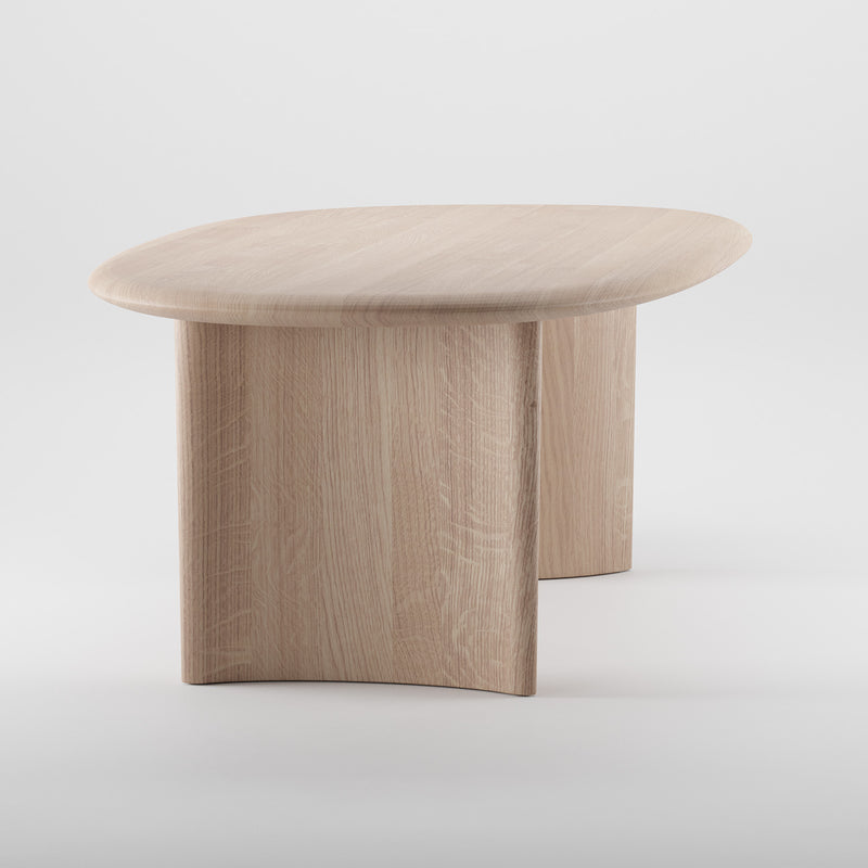 The MONUMENT Table by Artisan is a light wooden coffee table with a smooth oval top and a unique curved base. Made from solid wood with natural grain texture and finished in natural oils, it offers an eco-friendly, minimalist, modern look against a plain white background.
