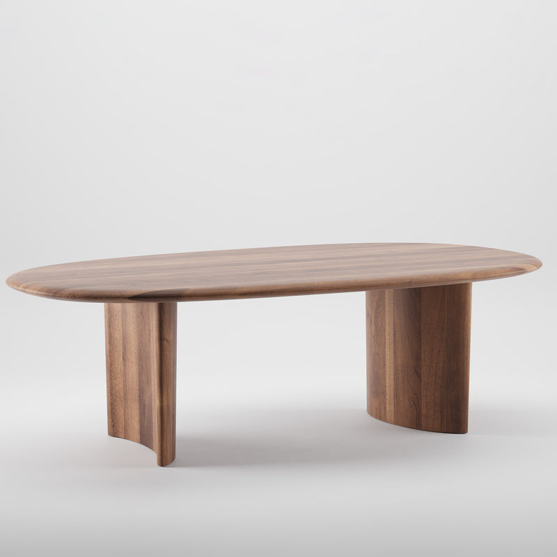 The Artisan MONUMENT Table is an oval wooden table with a polished surface and natural oils, featuring two sturdy curved legs. Crafted from solid wood using eco-friendly materials, its minimalist design stands elegantly against a plain white background.