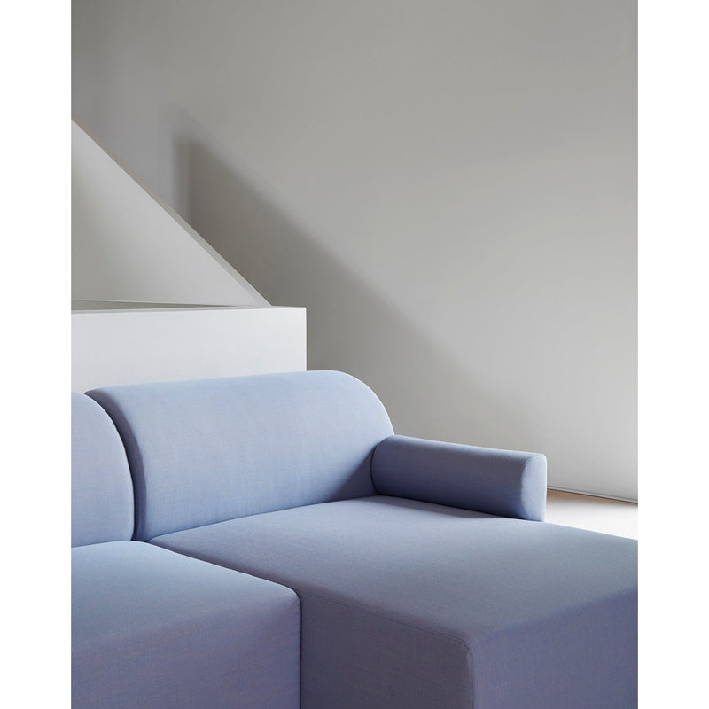 A minimalist room features a light gray wall with a geometric design. An Objekte unserer Tage Weber Modular Sofa in light blue, showcasing German craftsmanship and equipped with a cylindrical cushion on the armrest, sits in the lower left corner. Soft natural light casts subtle shadows.