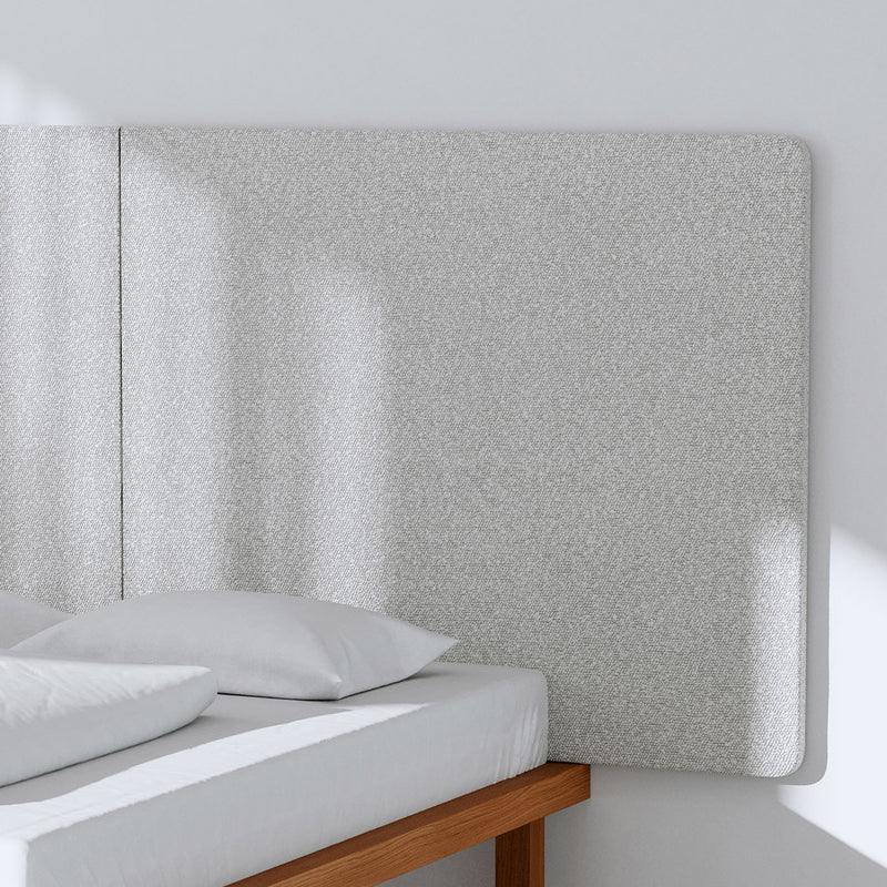 In a minimalist bedroom, a simple wooden bed frame holds a white mattress and pillows. A textured Frey MAX Headboard by Objekte unserer Tage with rounded corners complements the light gray wall. Soft shadows hint at natural light from a nearby window, enhancing the rooms serene acoustics.