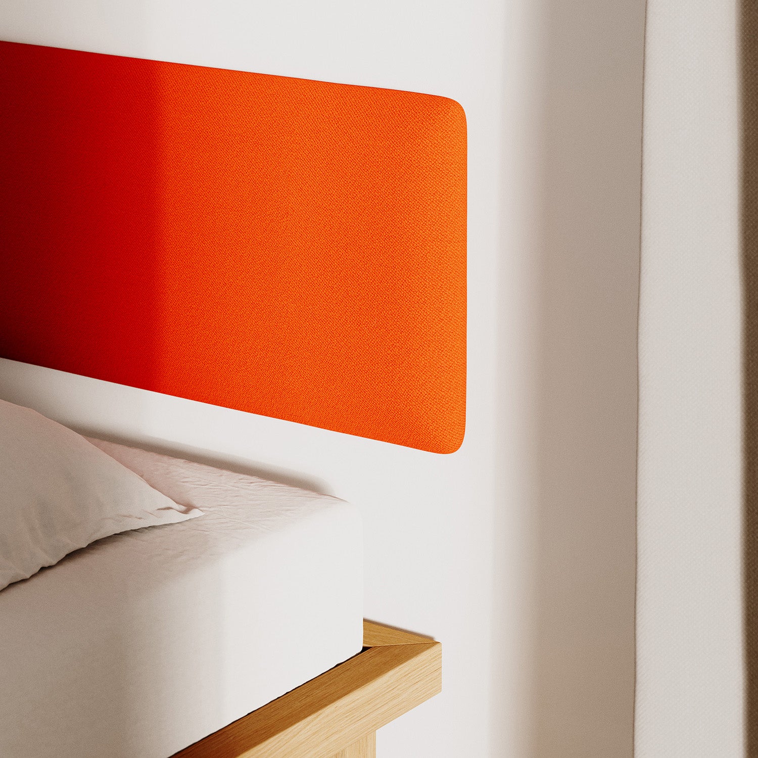 A cozy bedroom corner showcases a low wooden bed frame with a white mattress and pillow. Mounted on the white wall is the Frey Headboard by Objekte unserer Tage, crafted from bright orange fabric that casts a soft shadow. A flowing white curtain is partially visible.