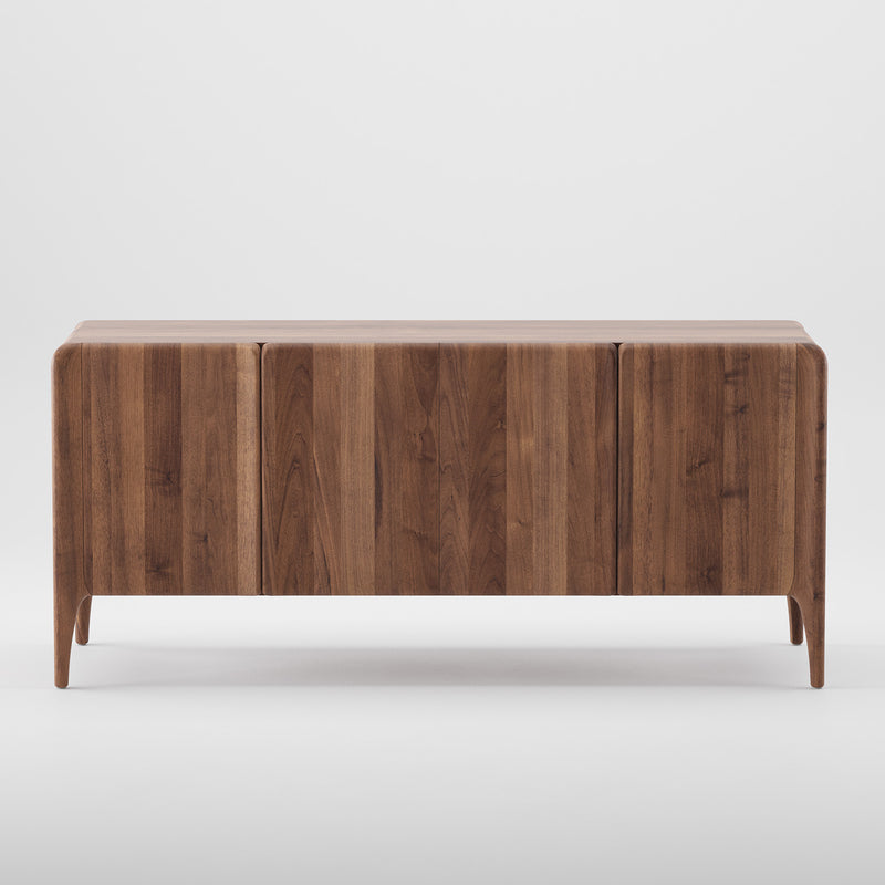 The RIO Sideboard by Artisan is crafted from solid wood with a sleek, minimalist design. It features three doors with subtle grain patterns, four narrow legs, and a smooth finish of natural oils and wax that beautifully complements a plain white background.
