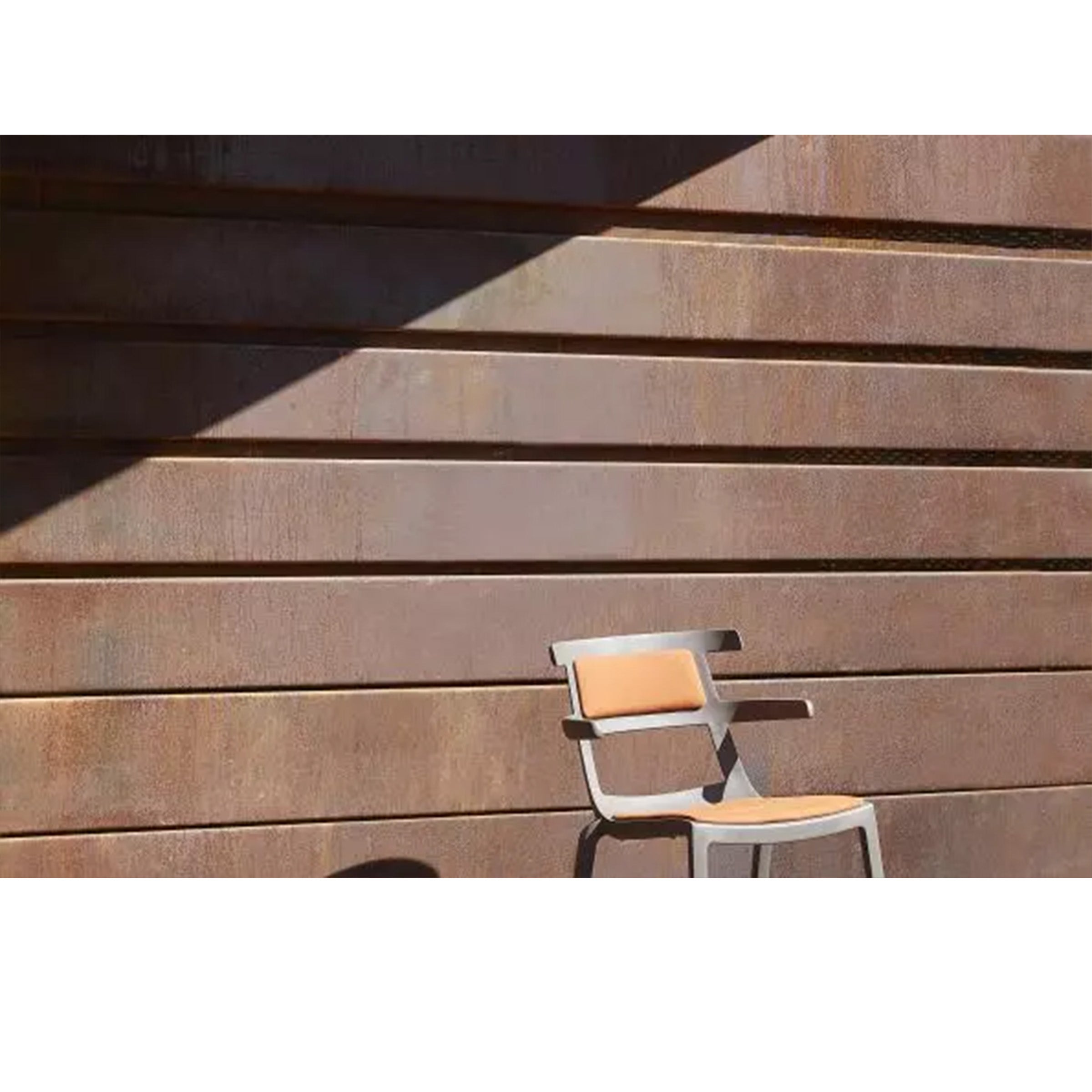 The Tokyo Chair Upholstered by Resol, featuring a beige seat, is set against a rust corrugated metal wall. Its UV-protected materials make it ideal for both indoor and outdoor use. A sharp diagonal shadow contrasts beautifully with the walls horizontal lines.