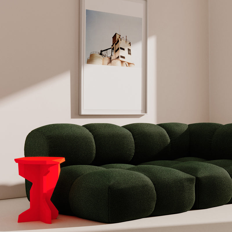 In a modern living room, a dark green, plush, tufted Sander Sofa from Objekte unserer Tage with ultrasoft upholstery is featured. Beside it sits a vibrant red table. A framed architectural photograph on the white wall adds to the minimalist look as shadows elegantly fall across the scene.