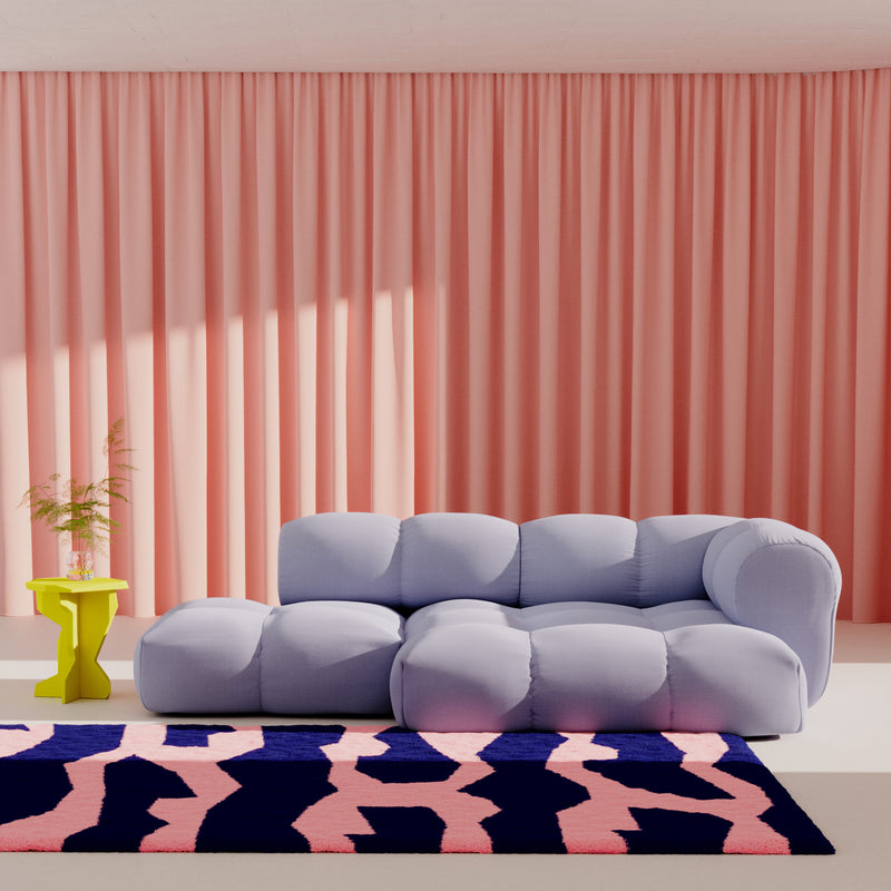 A modern living room features the light blue, tufted Sander Sofa by Objekte unserer Tage with its matching ottoman. Pink curtains serve as a backdrop, contrasted by a yellow side table with a plant. A bold pink and navy rug ties it all together, showcasing postmodernism elements.