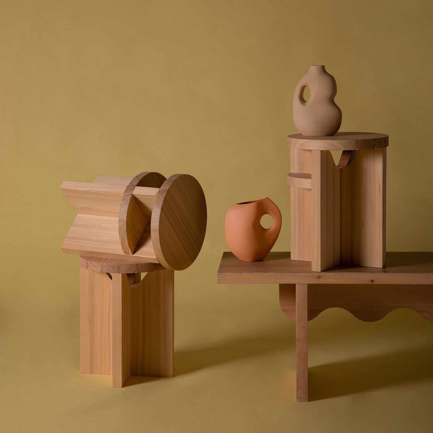 The Tami Dining Collection, featuring the Tami Stool by Schneid, showcases geometric wooden sculptures and clay vases in naturally oiled larch wood. Set on a mustard yellow background, these pieces highlight both curved and angular shapes for a modern abstract look.