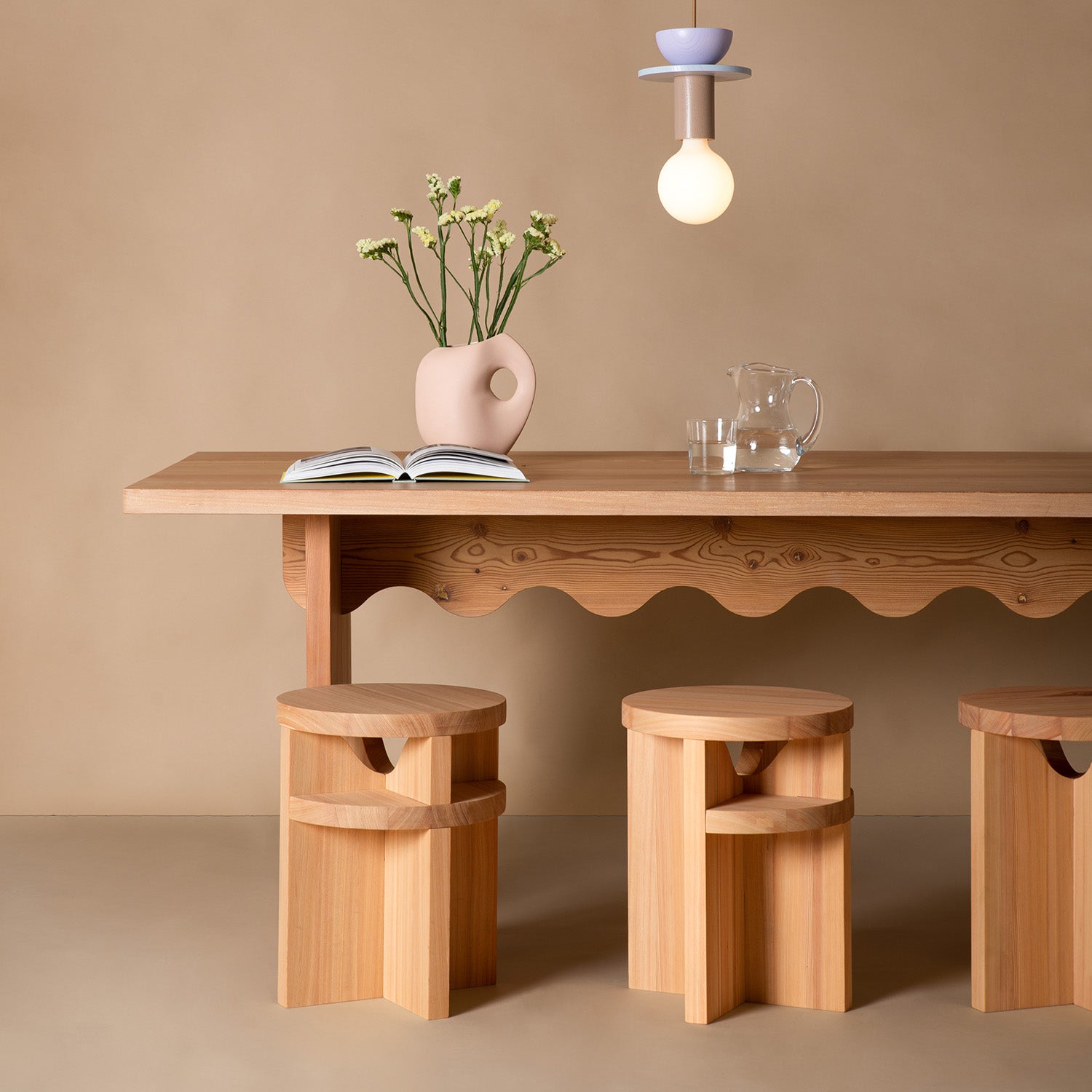 The Tami Dining Collection by Schneid includes a larch wood table complemented with a ceramic vase of white flowers, an open book, and a clear glass pitcher. Three Tami Stools fit perfectly underneath, while a modern light fixture hangs elegantly above against a beige backdrop.