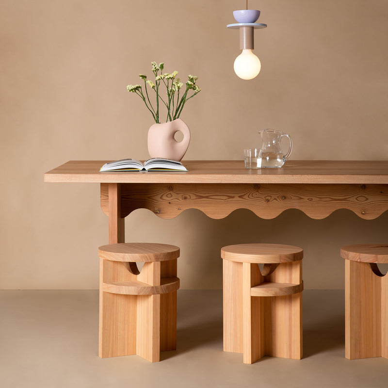 The Tami Dining Collection by Schneid includes a larch wood table complemented with a ceramic vase of white flowers, an open book, and a clear glass pitcher. Three Tami Stools fit perfectly underneath, while a modern light fixture hangs elegantly above against a beige backdrop.