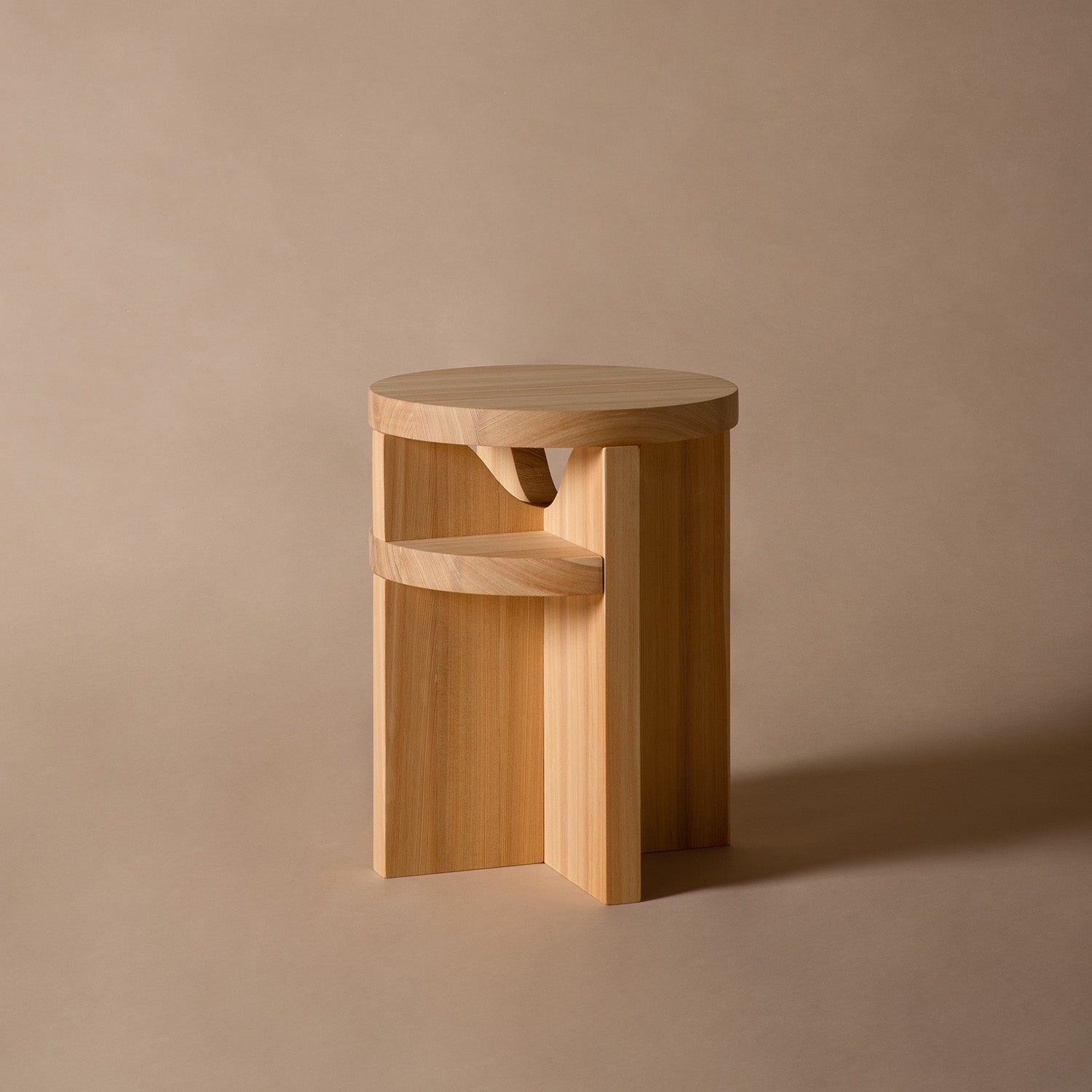 The Schneid Tami Stool, made from naturally oiled larch wood, showcases a round seat and interlocking geometric base. Positioned against a beige backdrop, it perfectly complements the refined style of the Tami Dining Collection.