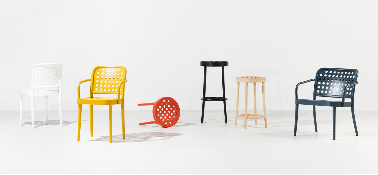 A minimalist scene with five modern chairs in an empty white room. The chairs are white, yellow, red, black, and blue, with the red chair lying on its back. Each chair has a distinctive, perforated design.