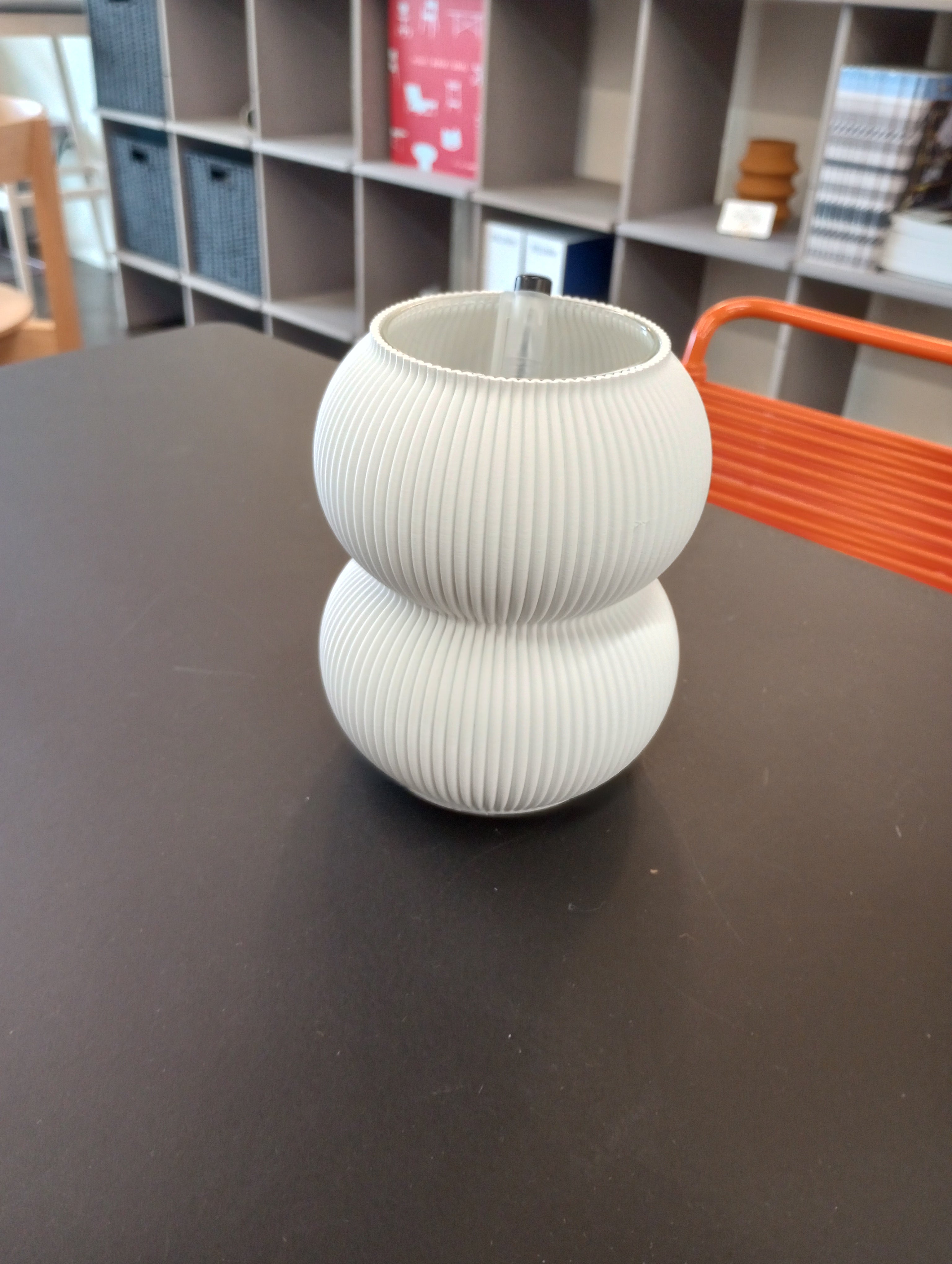 A Sheyn Zayl 159g Vase, with a ribbed, double-gourd shape and white finish, sits on a black table in the design studio. Shelves filled with books and storage boxes are in the background, along with the partial view of an orange chair.