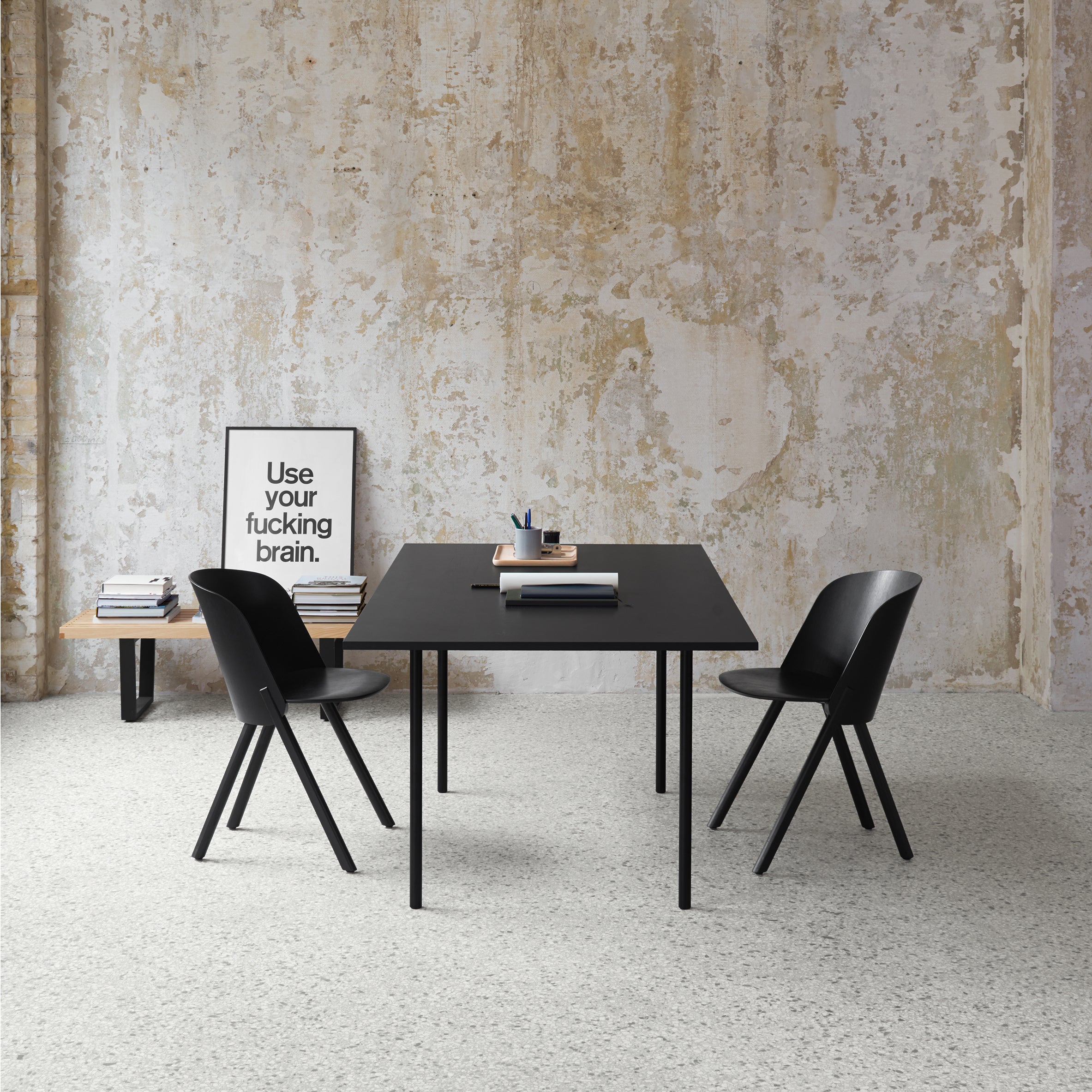 A minimalist room showcases the E15 TA14 ANTON, a sleek black table with steel legs, paired with two matching chairs on a speckled floor. A wooden bench displays books and a framed print. The textured peeling paint on the wall adds rustic charm, enhancing the overall style.