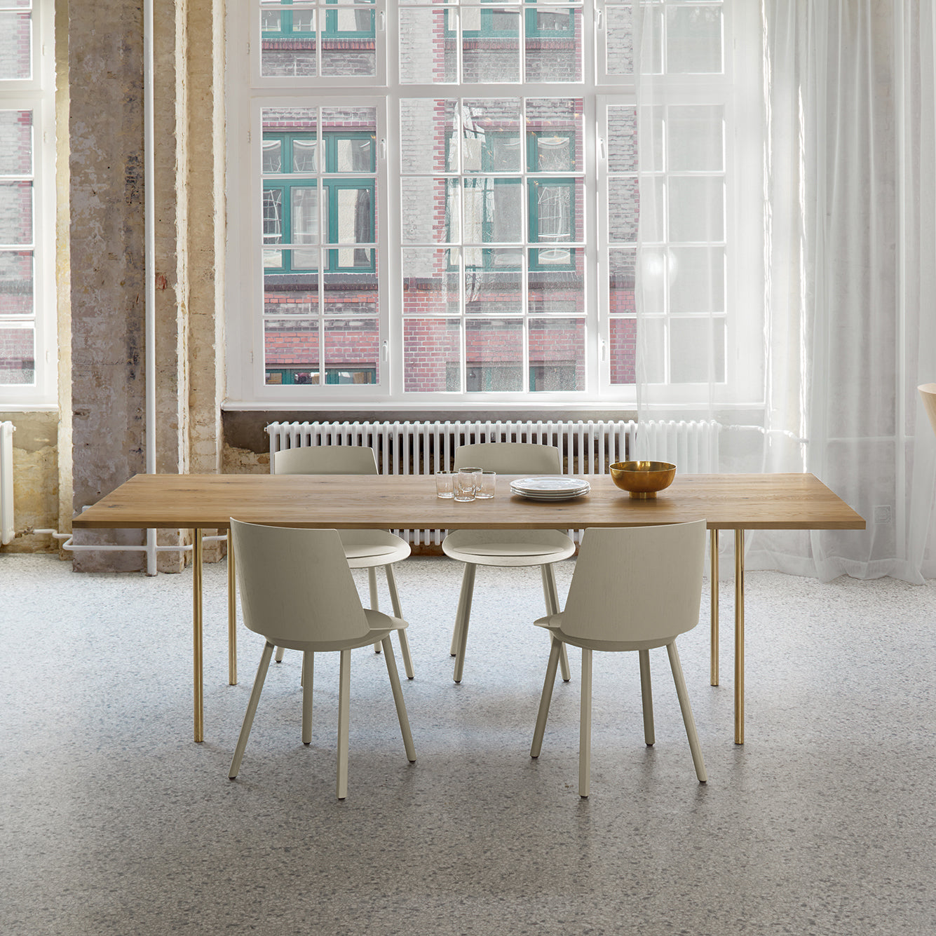 A minimalistic dining room showcases the TA14 ANTON solid wood table from E15, accompanied by four pale-colored chairs. On the table sit a glass carafe, cups, and a bowl. Large windows allow natural light to highlight a view of nearby brick buildings.