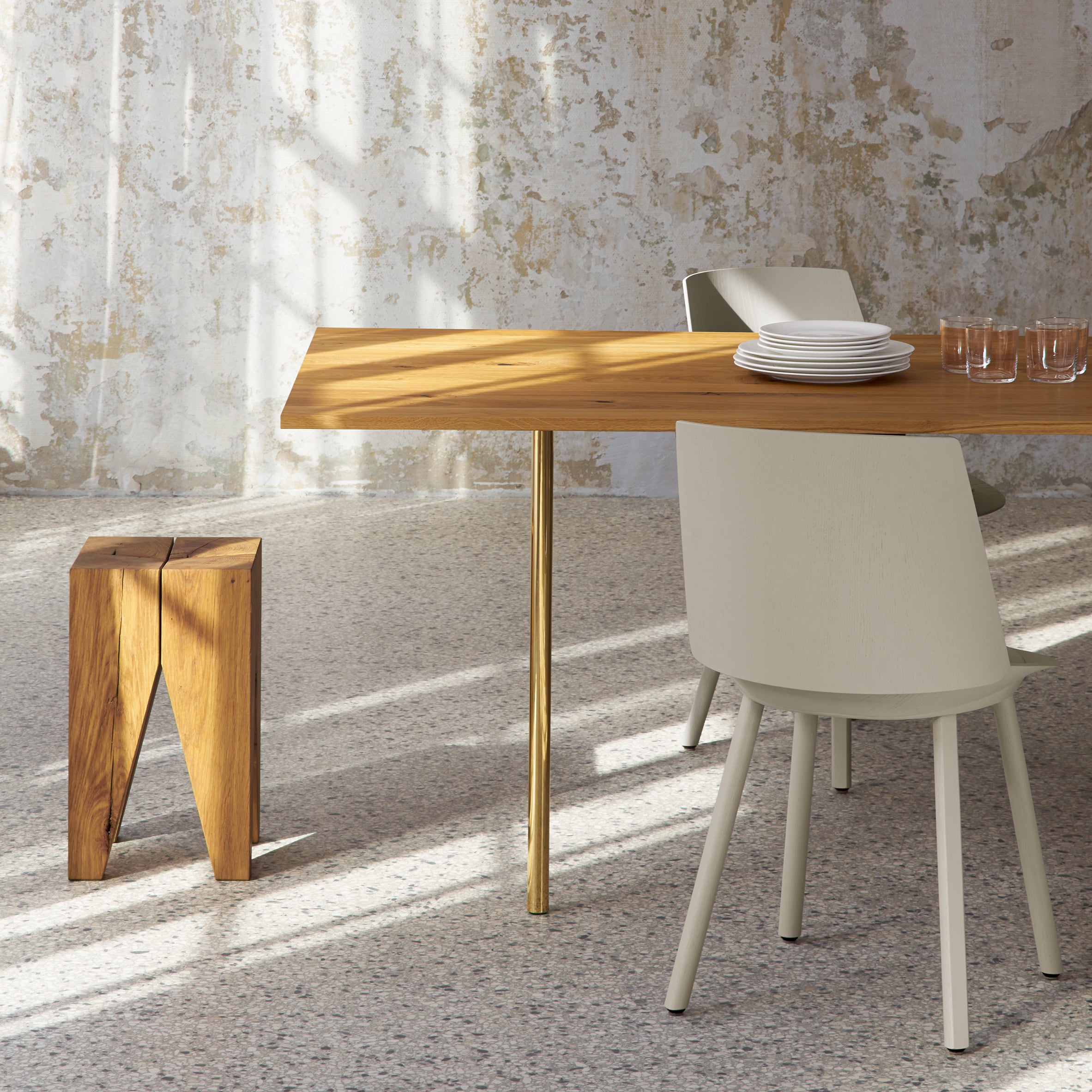 A modern dining area features the E15 TA14 ANTON table with a gold leg. Neatly stacked white plates and clear glasses sit on the walnut surface. Nearby, a wooden stool and beige chair enhance the setting as sunlight highlights intriguing shadows on the textured wall and floor.