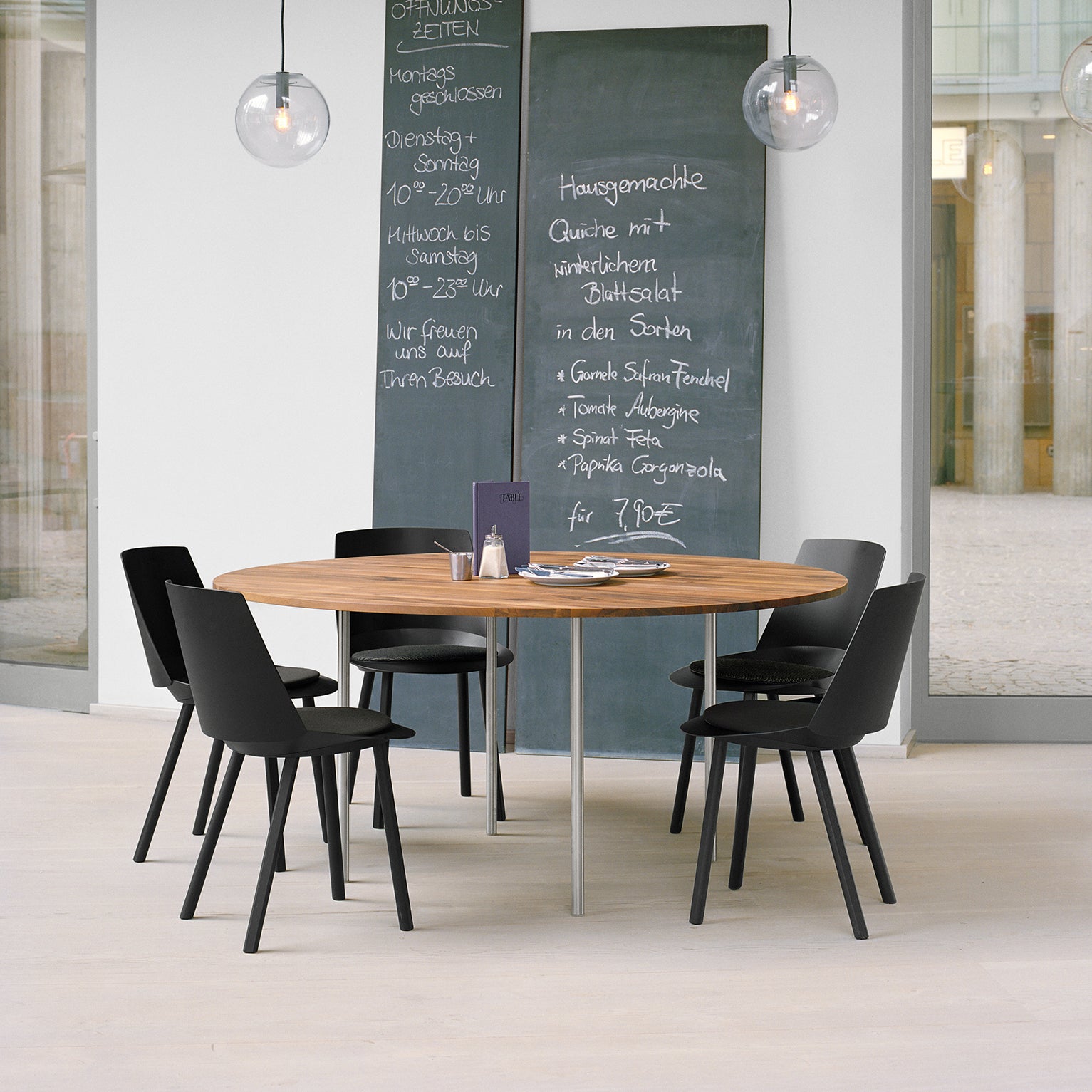 A modern dining area boasts the E15 TA14 ANTON round walnut tabletop paired with five black chairs. Two chalkboards show menu items in German, and globe lights brighten the minimalist space, highlighting large windows and a sleek solid wood table with steel legs.