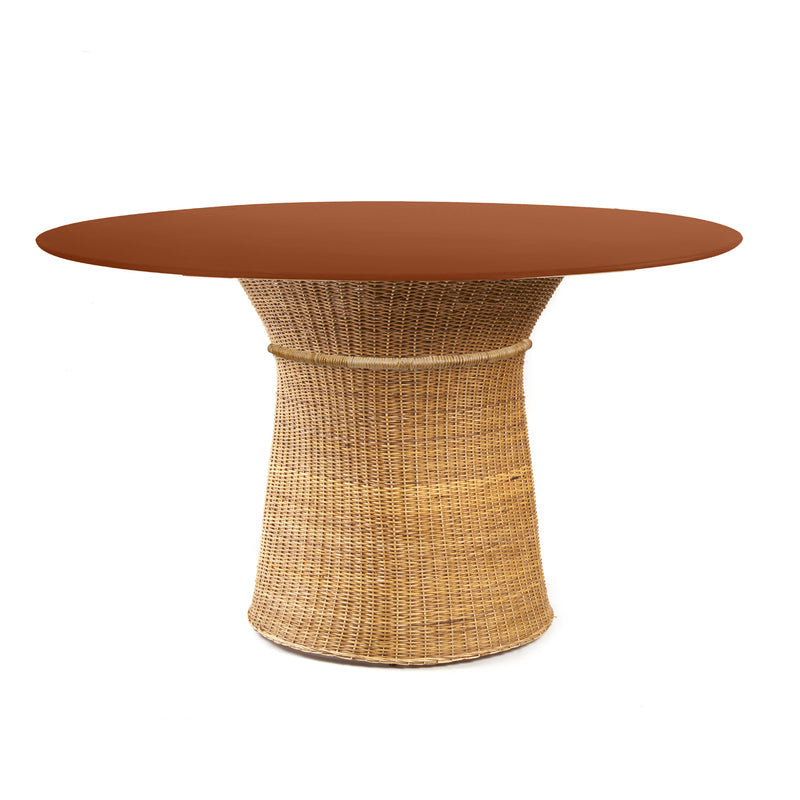 The Ames Caribe Natural Dining Table by Ames features a round, smooth reddish-brown top and a natural wicker base that flares out at the bottom, photographed against a white background—an elegant piece inspired by Sebastian Herkner’s designs.