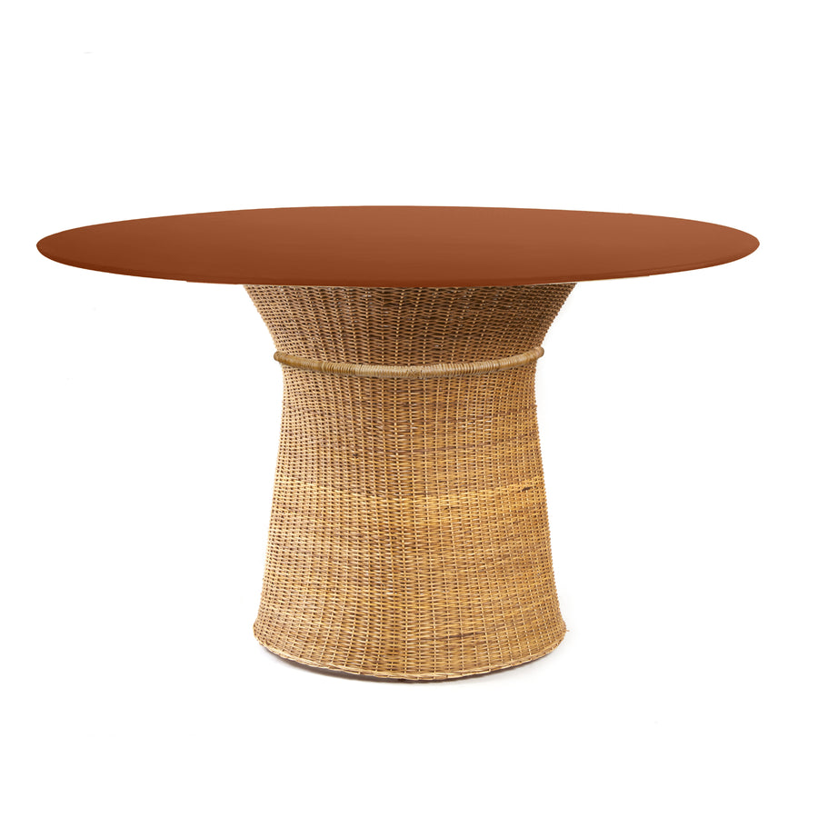 The Ames Caribe Natural Dining Table by Ames features a round, smooth reddish-brown top and a natural wicker base that flares out at the bottom, photographed against a white background—an elegant piece inspired by Sebastian Herkner’s designs.
