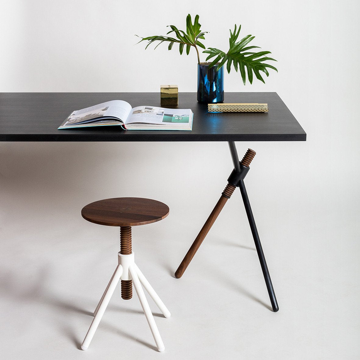 A modern black table holds a plant in a blue vase and an open magazine. Nearby is the Thread Family - Stool by Coordination Berlin, featuring a round walnut wood seat and tripod legs with large screw-like designs for customizable colors.