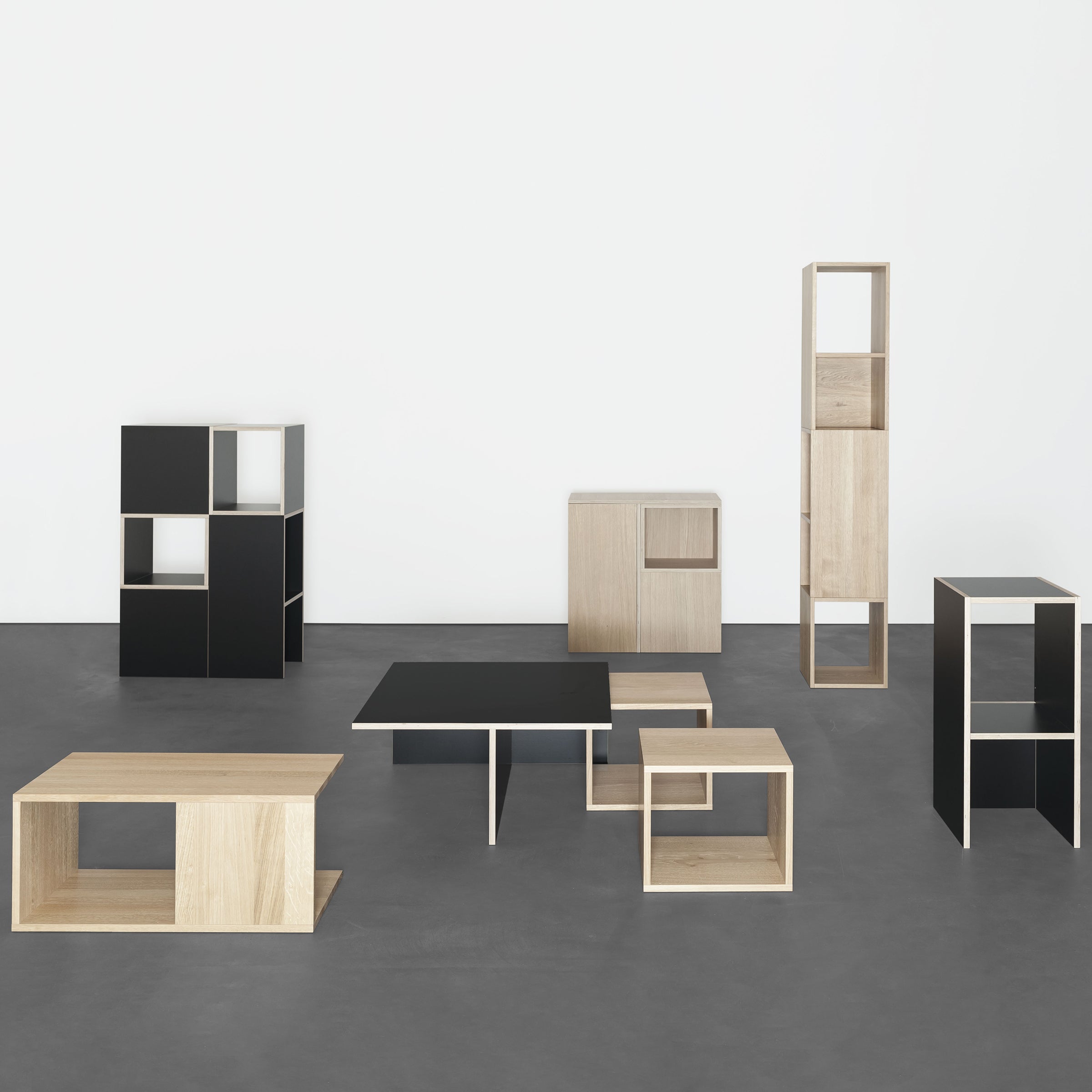 A minimalist space features Sanktjohansers Basic multipurpose modular furniture, showcasing geometric wooden pieces in black and natural finishes. The setup includes a low table, shelves, and customizable cubical storage units on a smooth gray floor against a plain white wall.