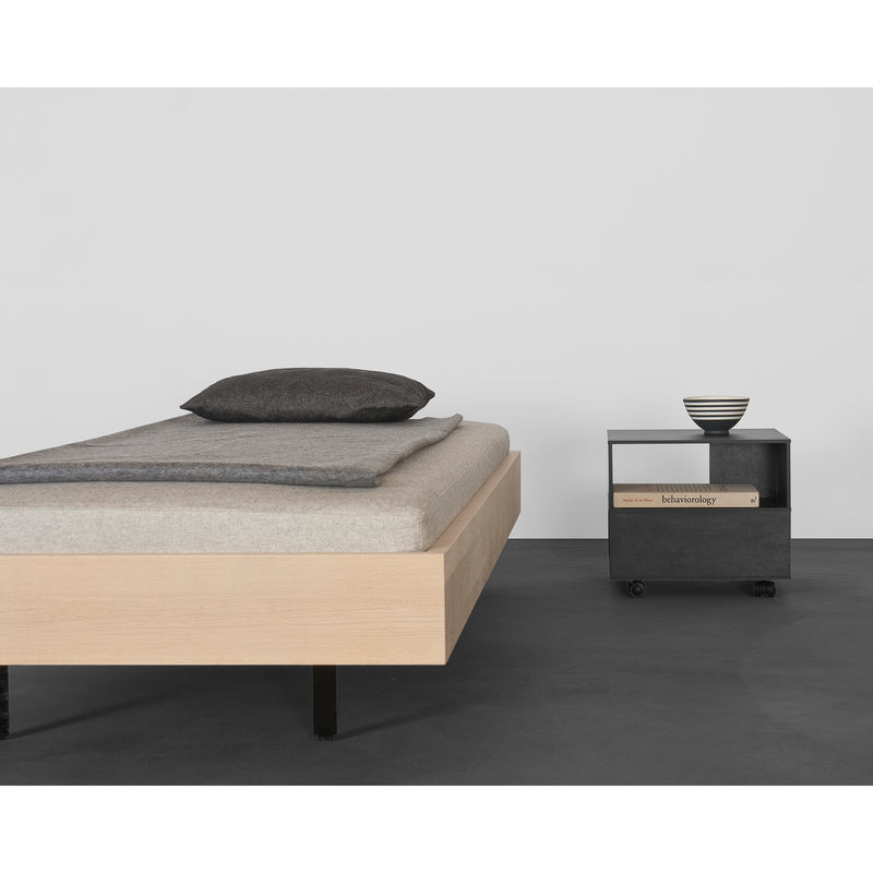 A minimalist bedroom features the Sanktjohanser Iku Bed Series, a solid wood platform with a gray mattress, pillow, and blanket. A small dark nightstand on wheels holds Kulturgeschichte and a striped bowl. The room has sleek gray flooring and pristine white walls.
