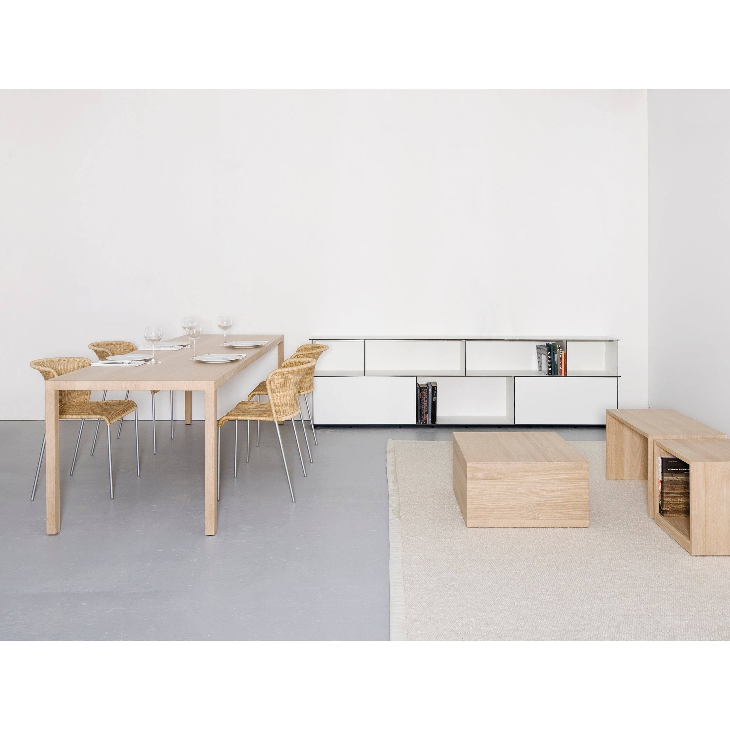 A minimalist living and dining space showcases a light wooden dining table with wicker chairs, set with glassware. A low wooden coffee table and bench rest on a beige rug. A modular sideboard backs the wall next to a white open-shelf unit with books. Featuring the Digital multipurpose by Sanktjohanser.