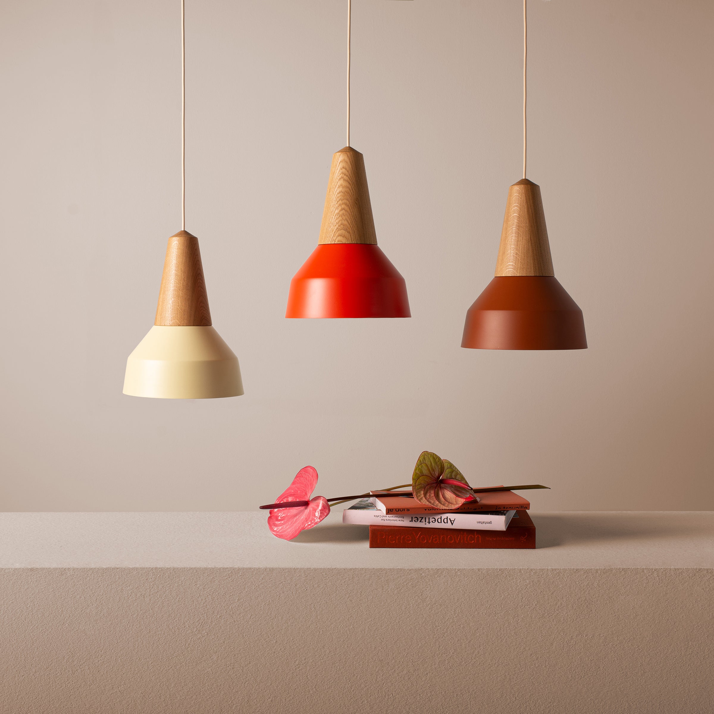 Three Eikon Basic Wax pendant lights from Schneid with elegant cream, red, and brown lampshades hang above a minimalist beige shelf. Two anthurium flowers enhance certified wood bookshelves holding three uniquely covered books against a neutral, soft-toned wall.