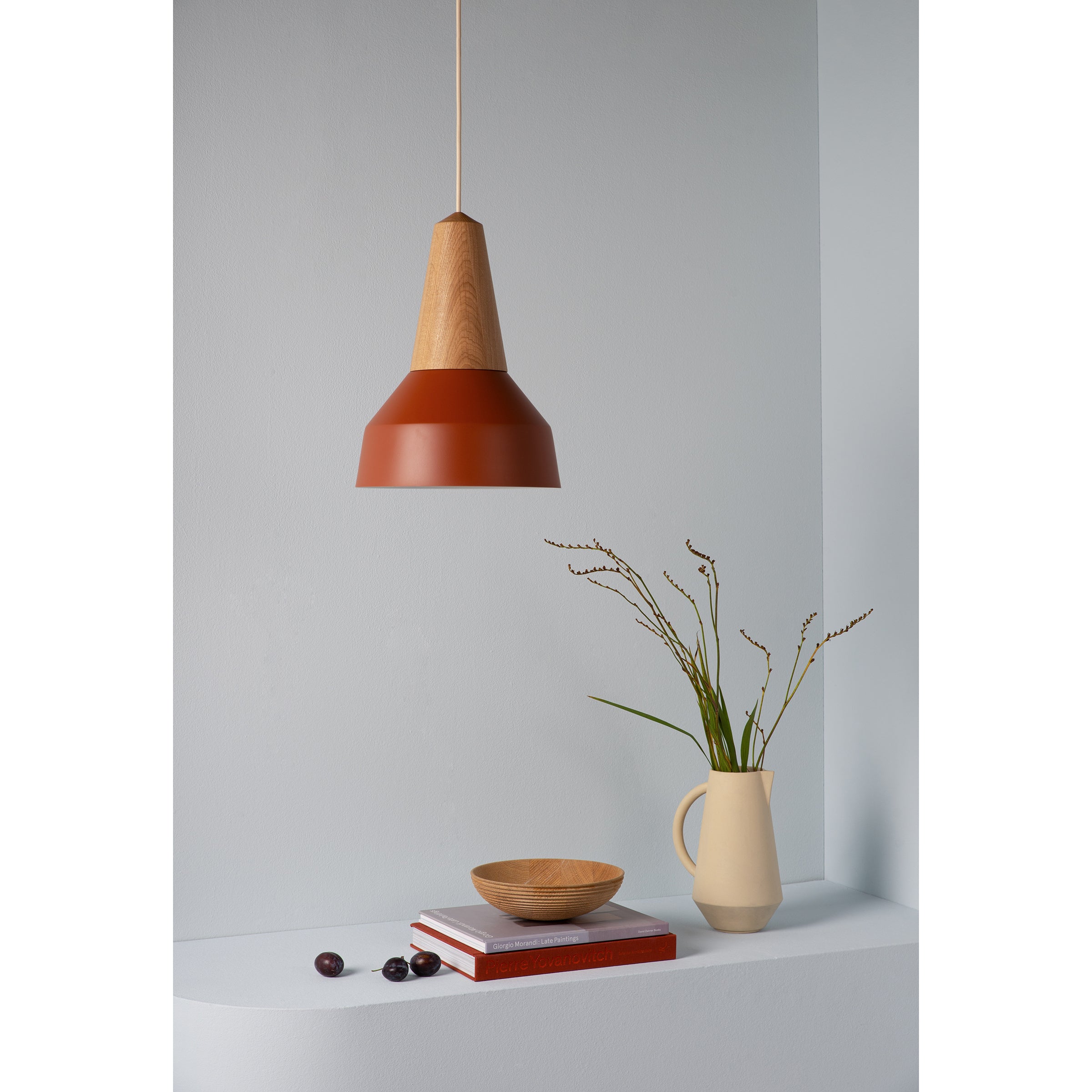 A modern room corner highlights a minimalist design with Schneids Eikon Basic Amber pendant lamp, a certified wood bowl on books, two cherries, and a beige vase with twigs on a white shelf against a pale blue wall.
