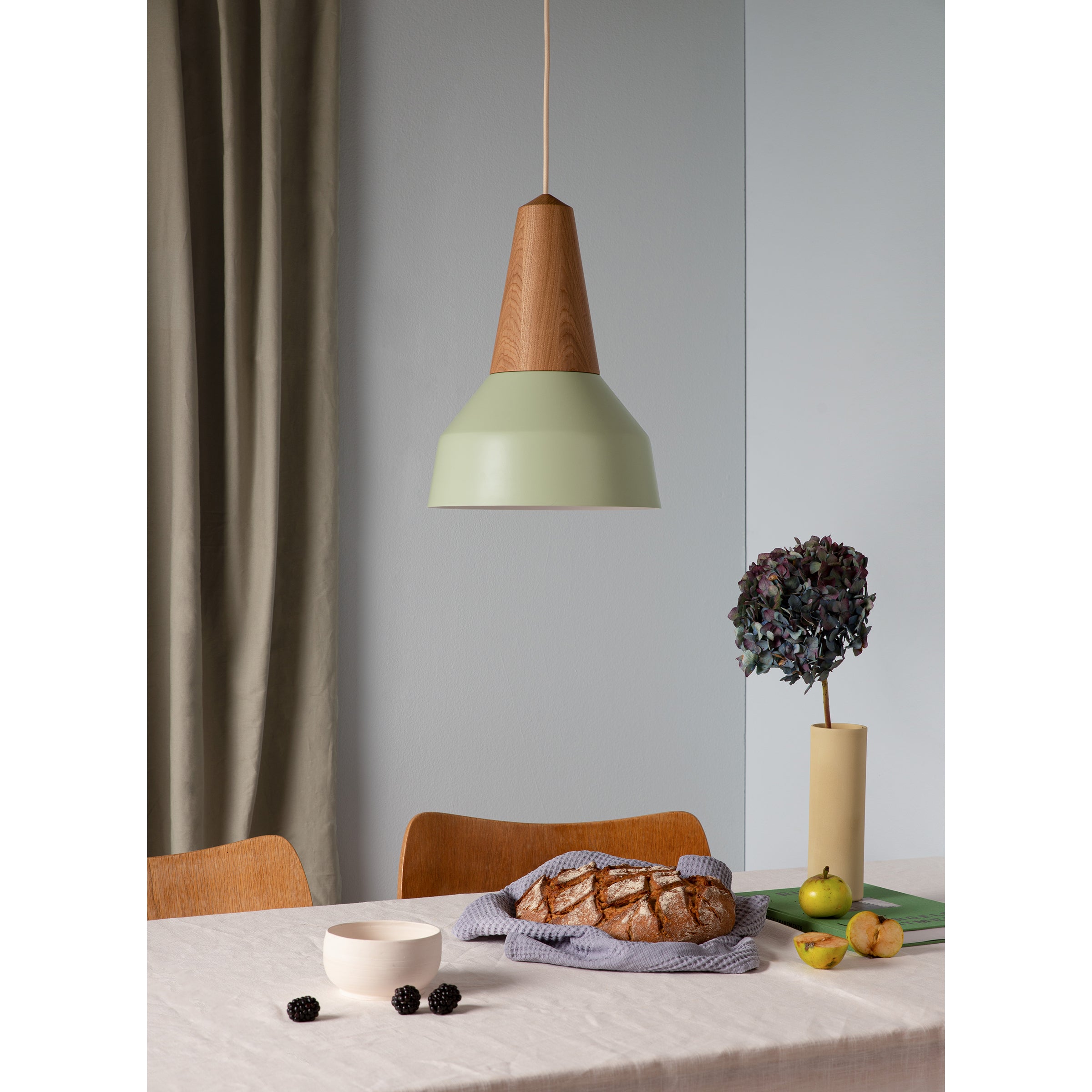 A modern dining area features a wooden table made from certified wood with a cloth, flanked by two wooden chairs. Above, a Schneid Eikon Basic Pistache lamp illuminates dried flowers and a spread of blackberries, rustic bread, apples, and pears.
